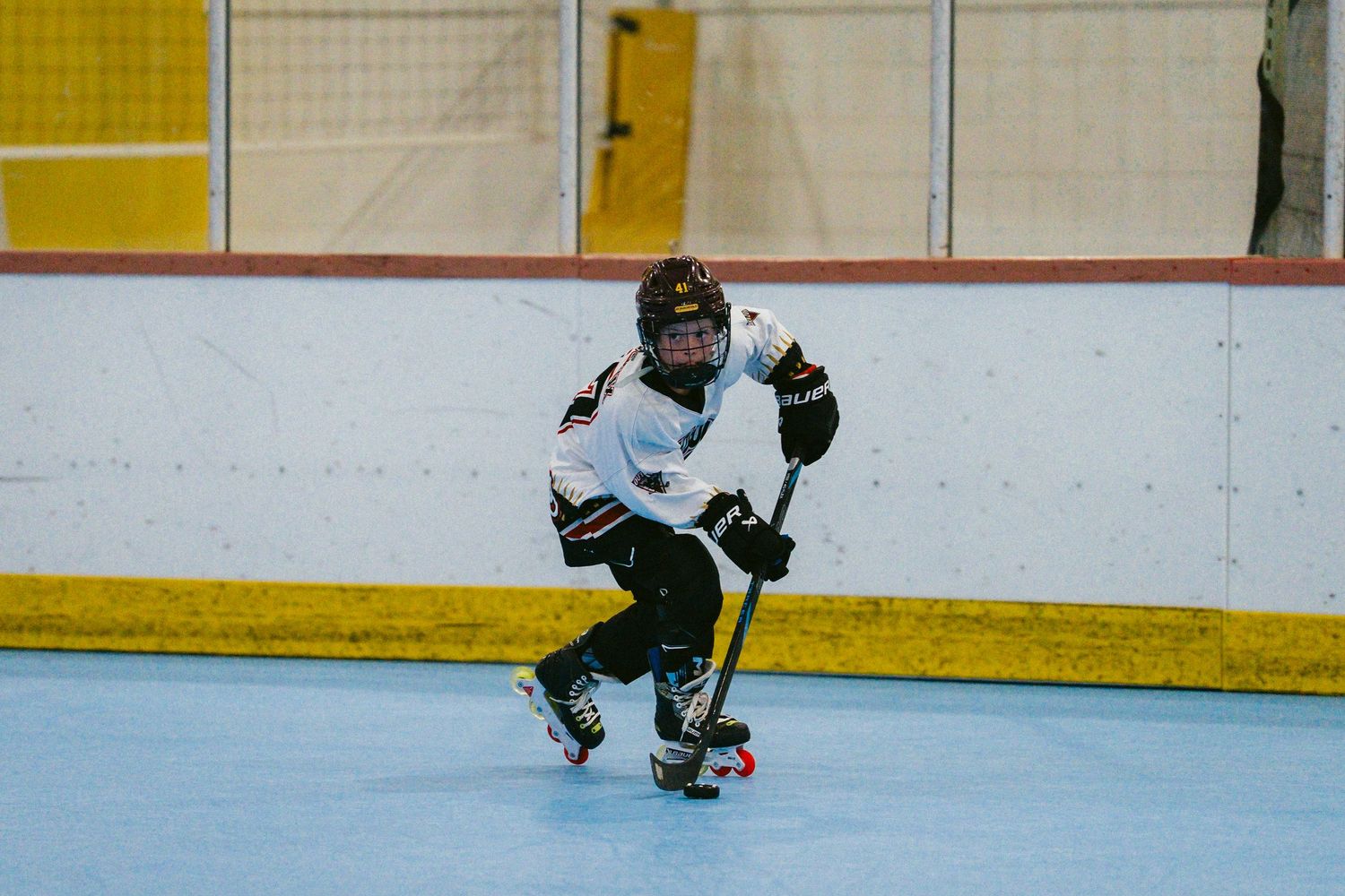Young hockey player skating on indoor rink in white and black uniform, holding stick during a game.