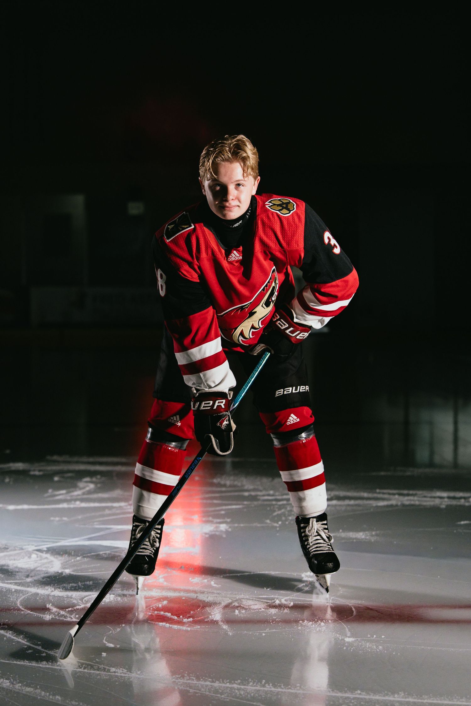 Young male hockey player in red Arizona Coyotes jersey posing with stick on ice rink.