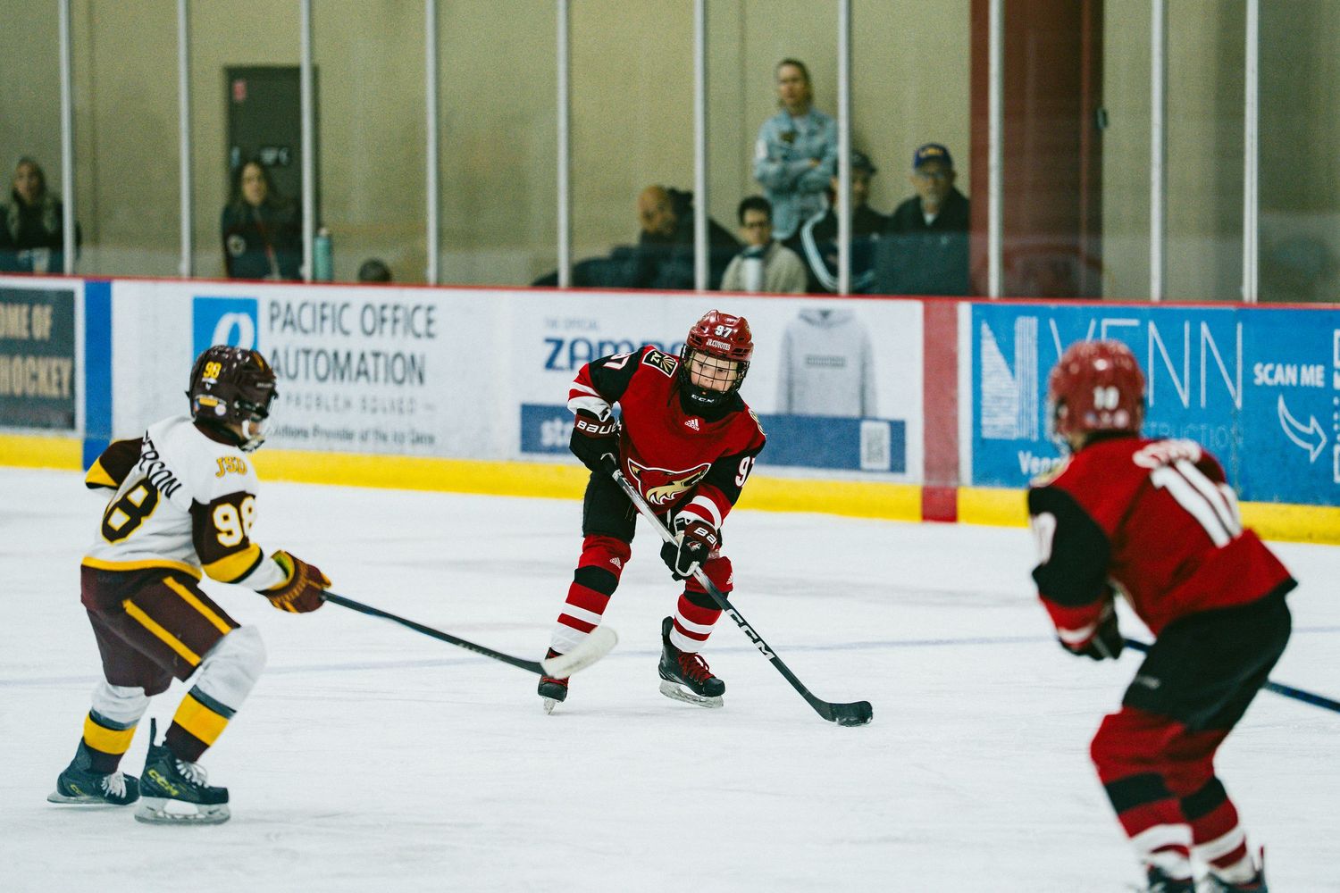 Hockey players in red and brown jerseys face off during an ice hockey game on a rink with advertising boards visible.