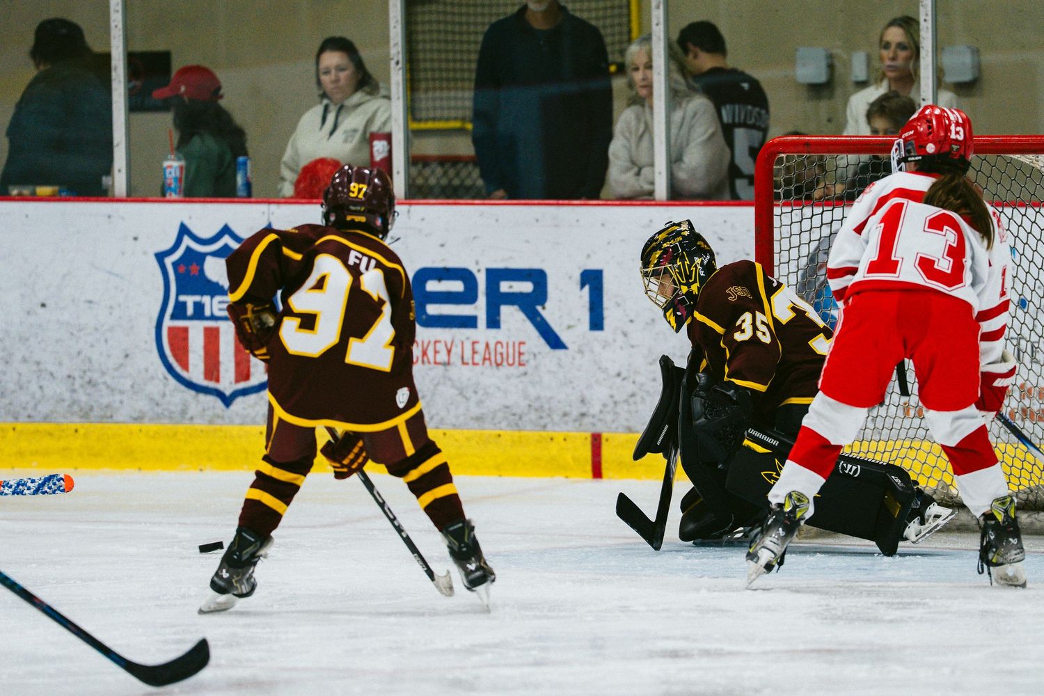 Youth hockey players in brown and red jerseys compete during an intense ice hockey match.
