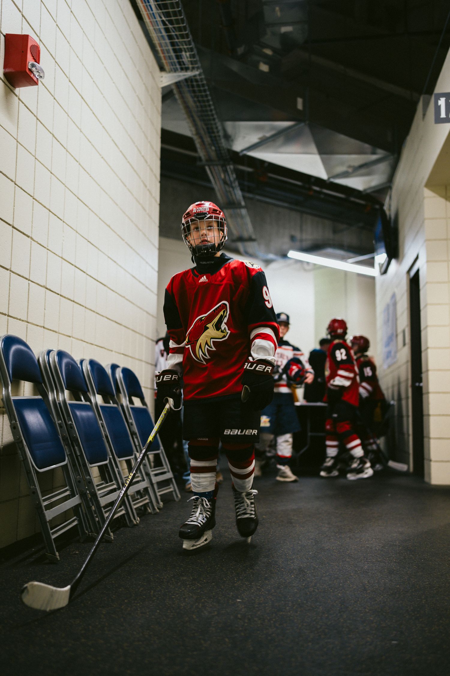 Hockey player in red Arizona Coyotes jersey stands in arena hallway near folding chairs.