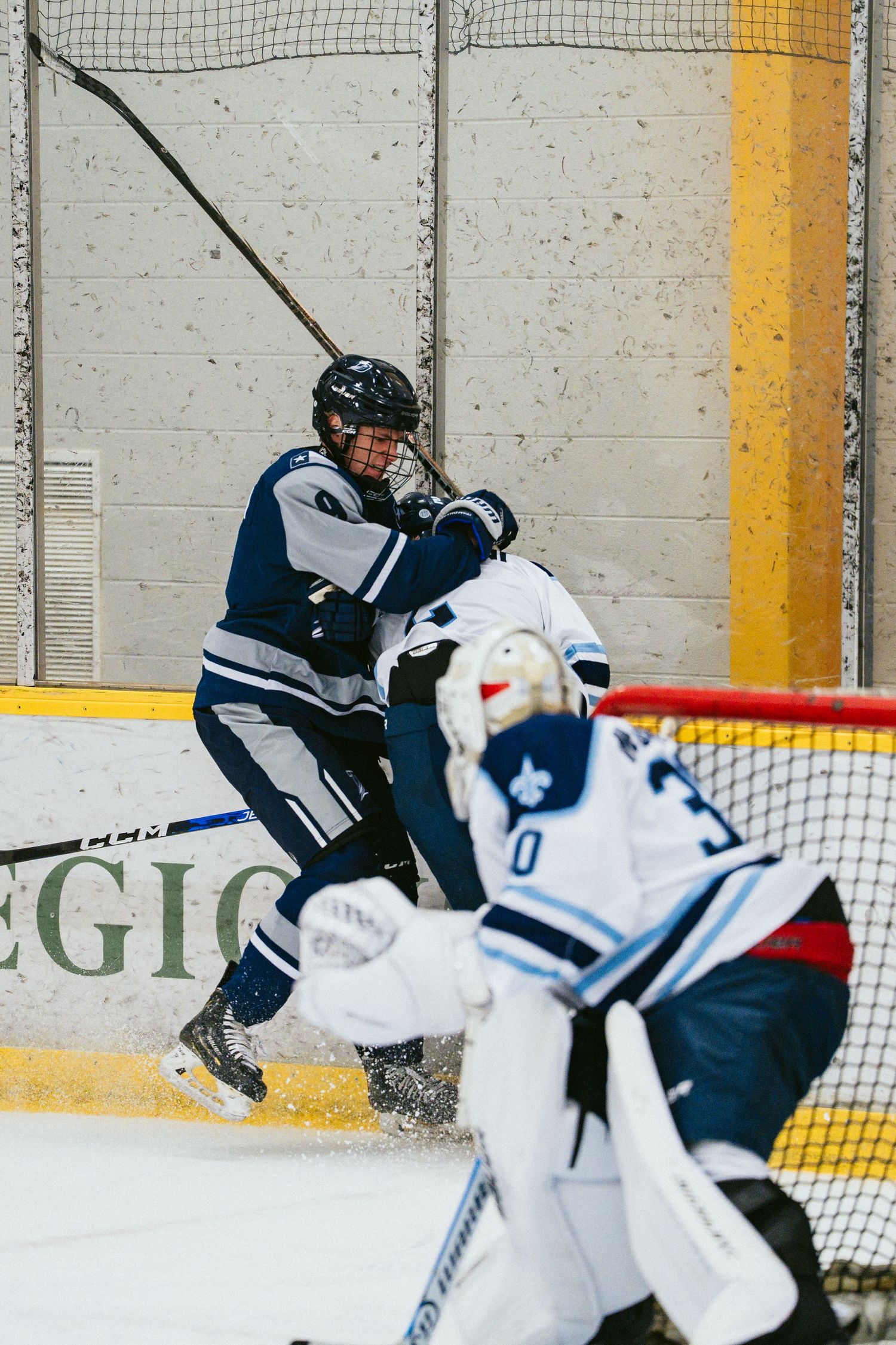 Two ice hockey players battle along the boards as a goalie watches during an intense hockey game.