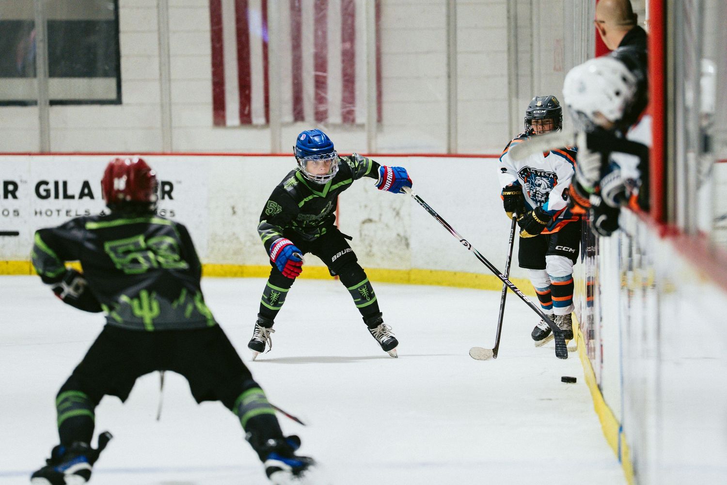 Youth ice hockey players in green uniforms defend against opponents during an indoor hockey game.