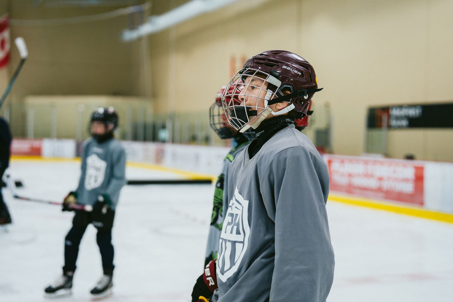 Young hockey player in maroon helmet and gray jersey stands on indoor ice rink during practice session.