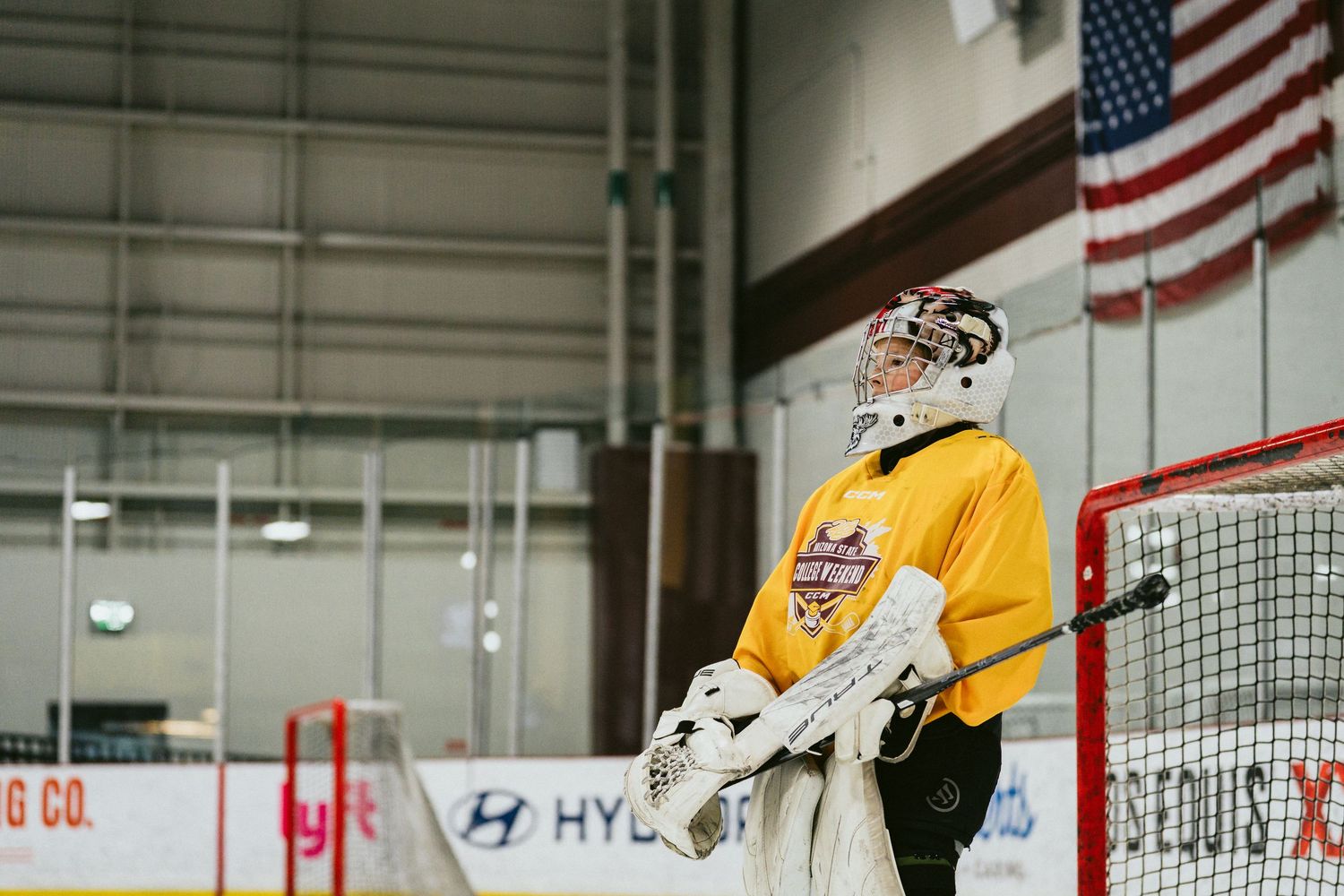 Hockey goalie in yellow jersey stands guard at net inside indoor ice rink with American flag.