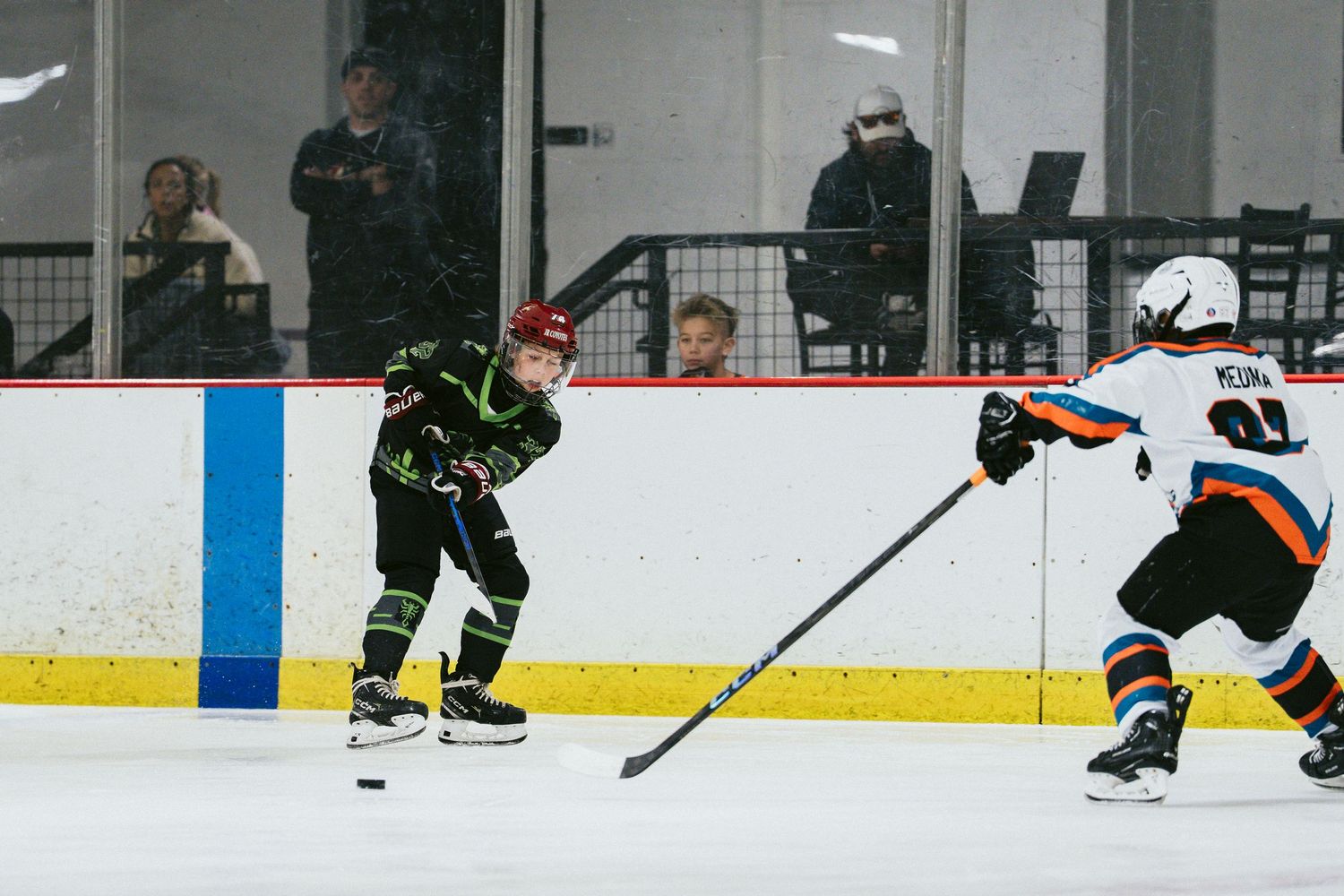 Youth hockey player in black green uniform prepares to pass puck against opponent in white jersey on ice.