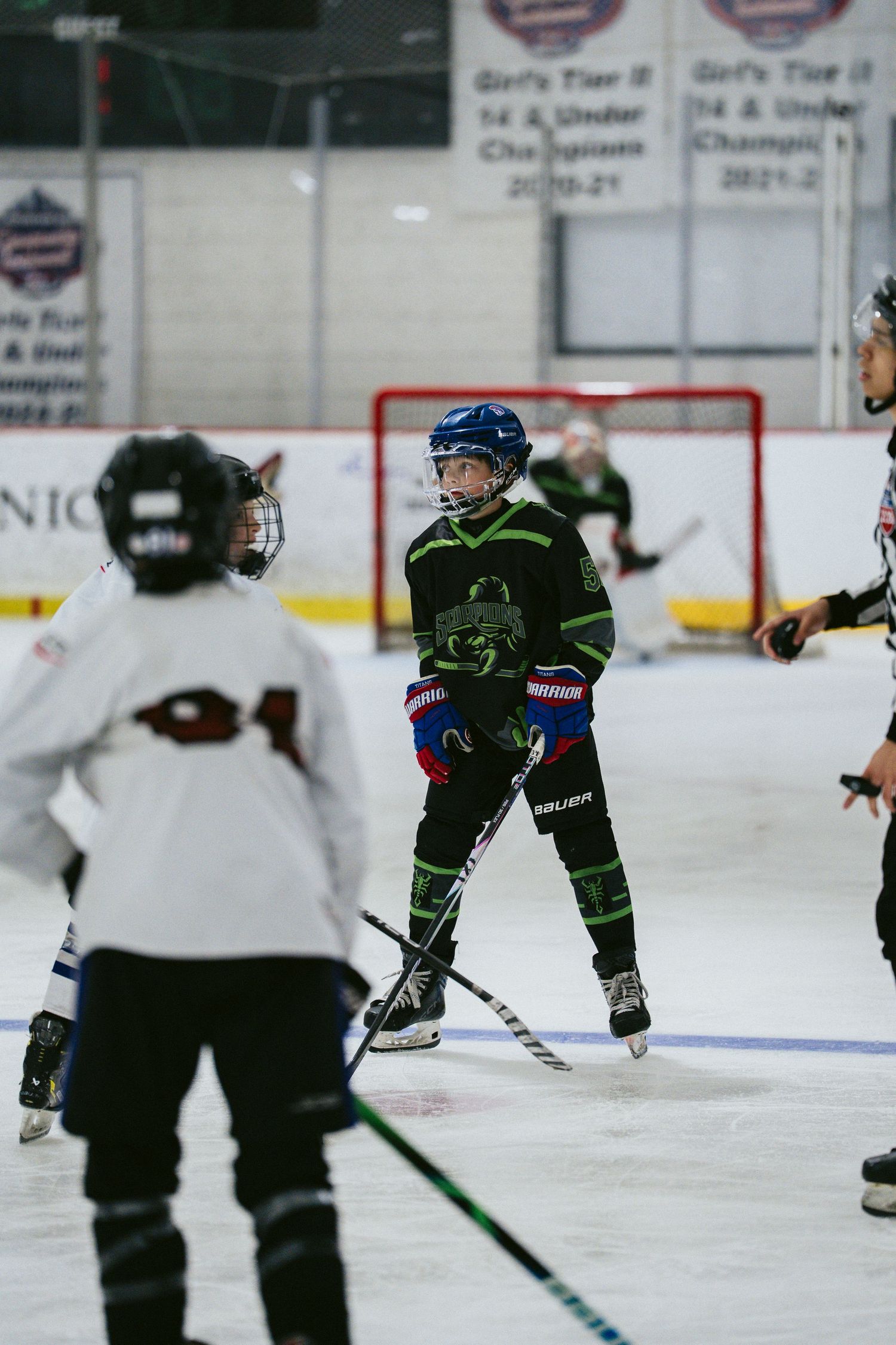 Youth ice hockey player in black and green uniform stands on ice during indoor hockey practice or game.