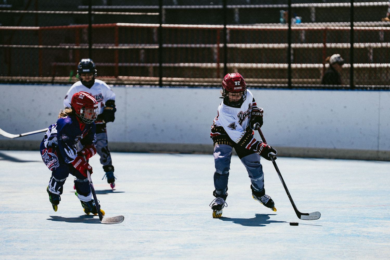 Young children playing roller hockey on an outdoor rink wearing helmets and protective gear.