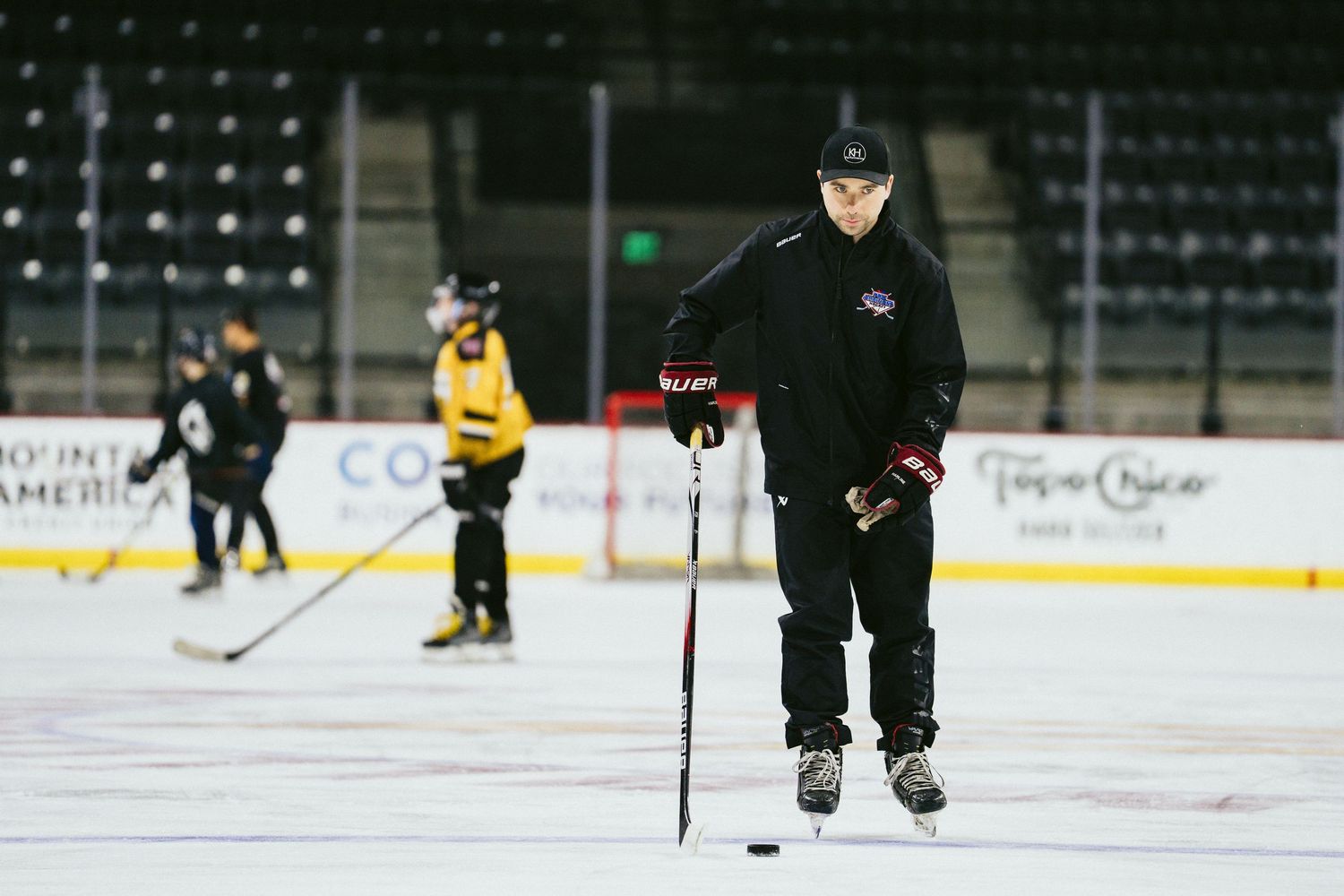 Ice hockey coach in black uniform stands with stick and puck during youth practice on indoor rink.