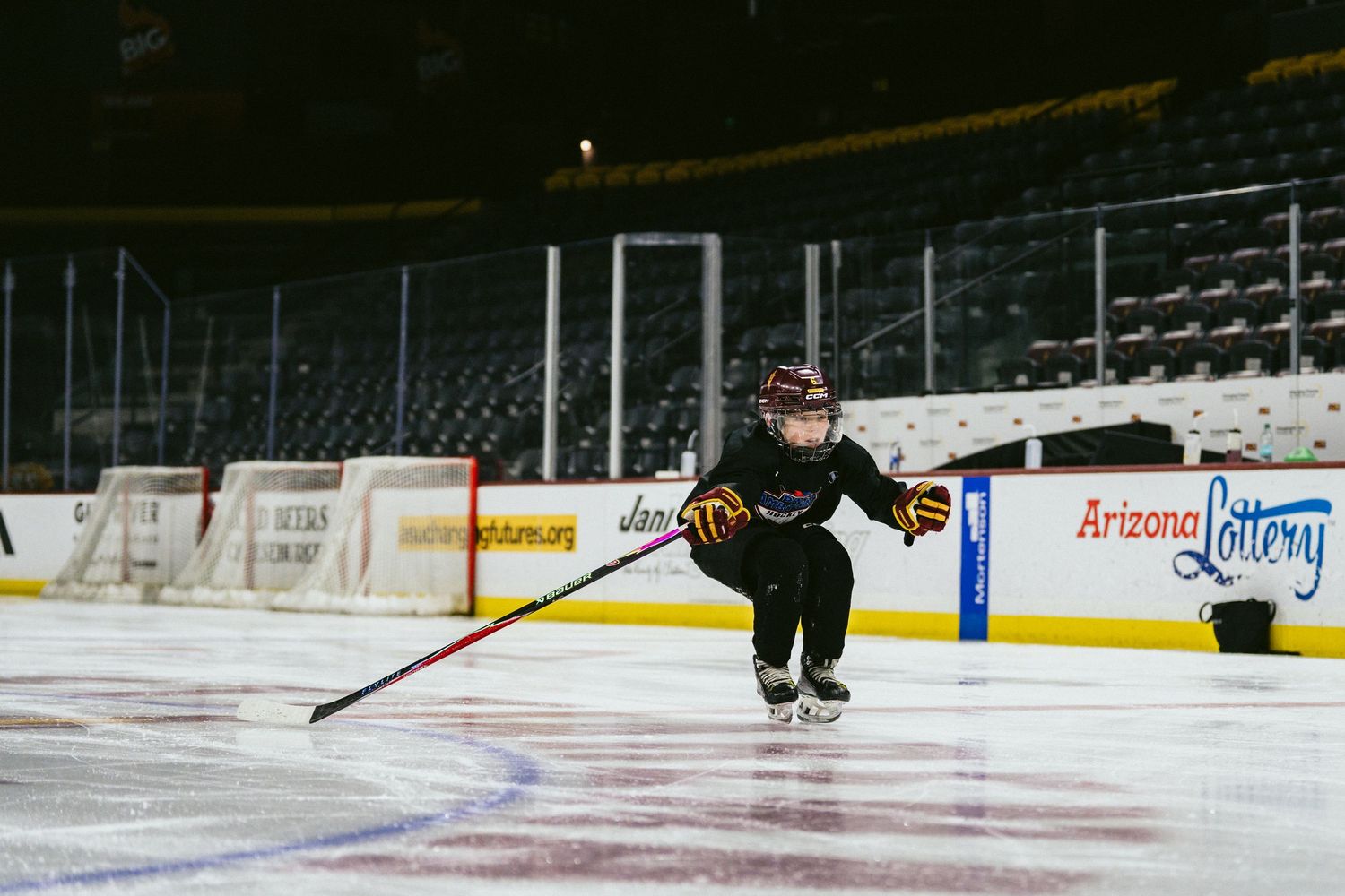 Young child skates on ice rink holding hockey stick during youth hockey practice at indoor arena.