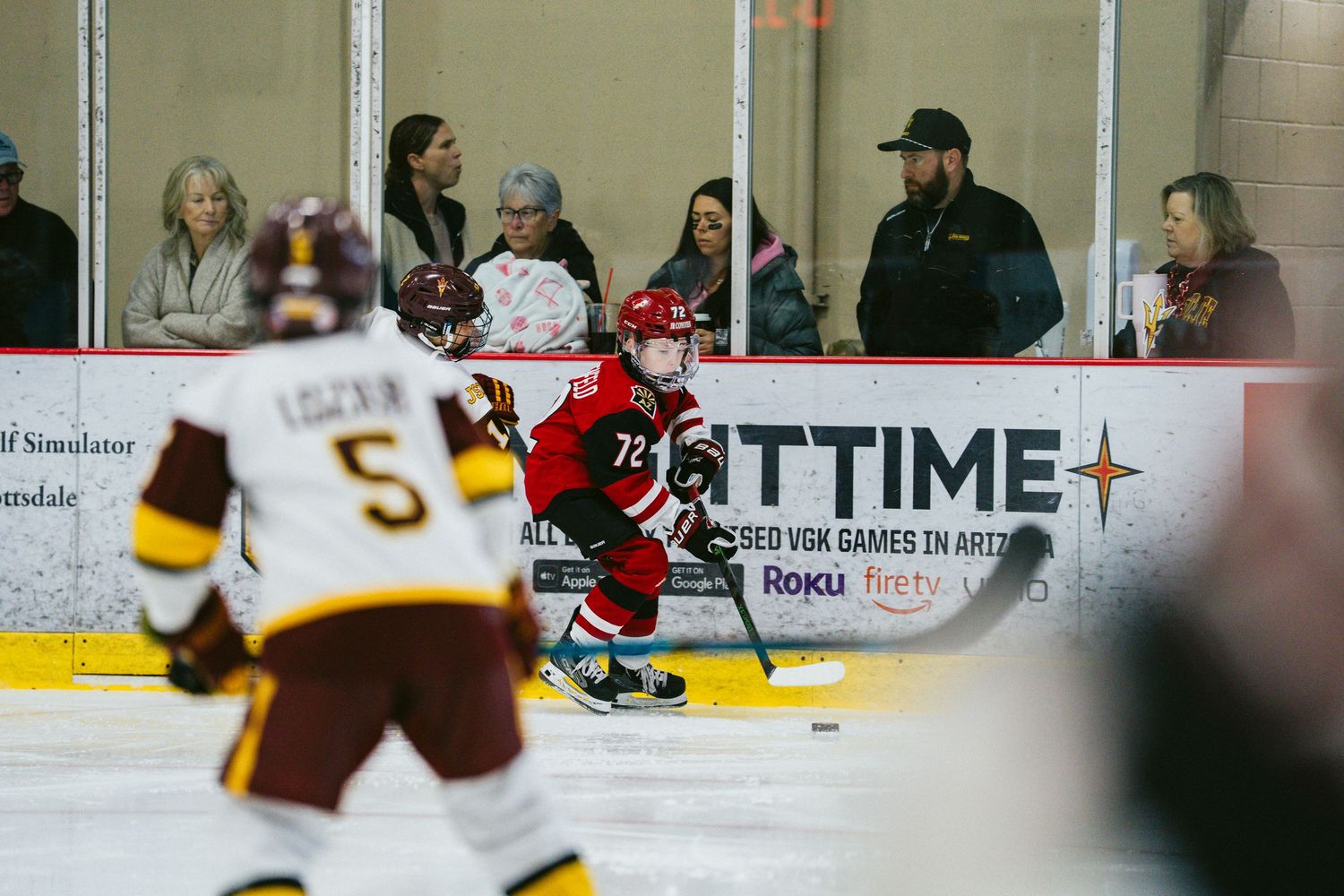Ice hockey players from opposing teams in red and brown uniforms compete during a match along the boards.