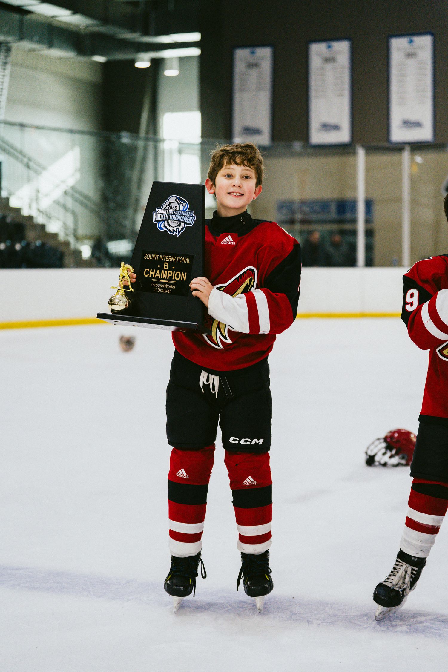 Young hockey player in red uniform proudly showing off championship trophy on ice rink.