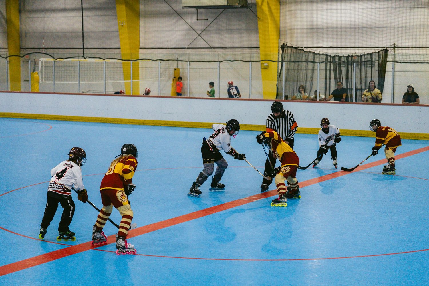 Young roller hockey players face off on a blue indoor rink with a referee overseeing the game.
