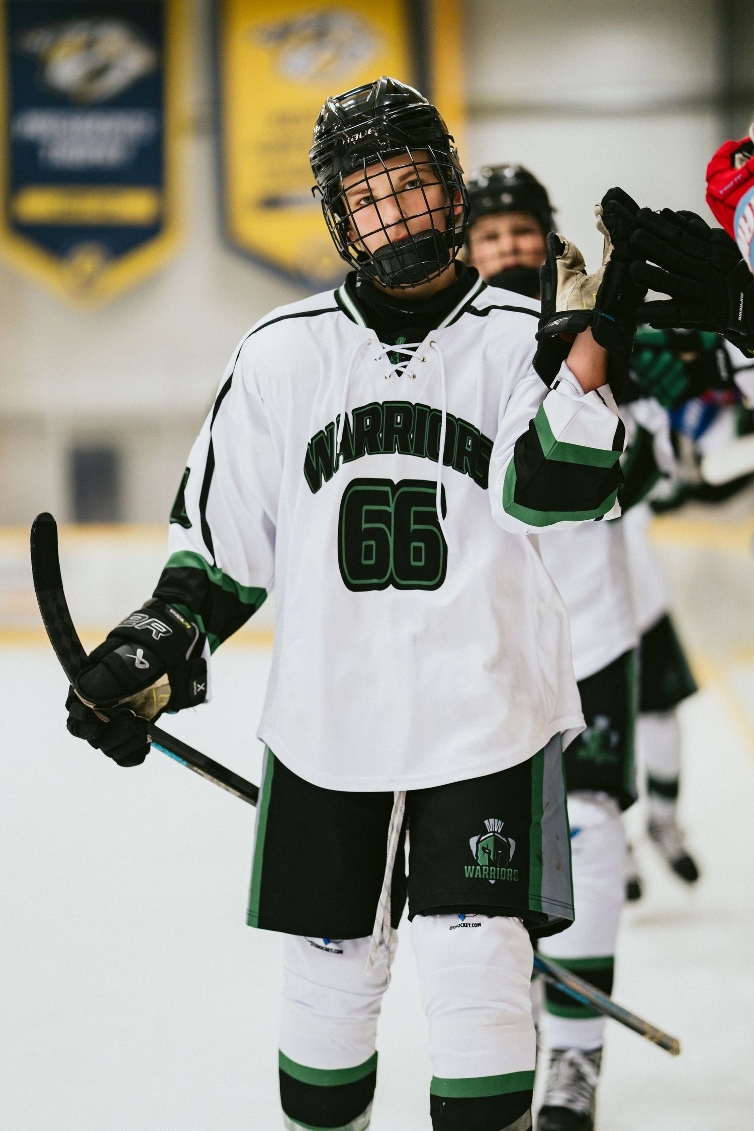 Youth hockey player wearing number 66 Warriors jersey holds stick on ice rink during game.
