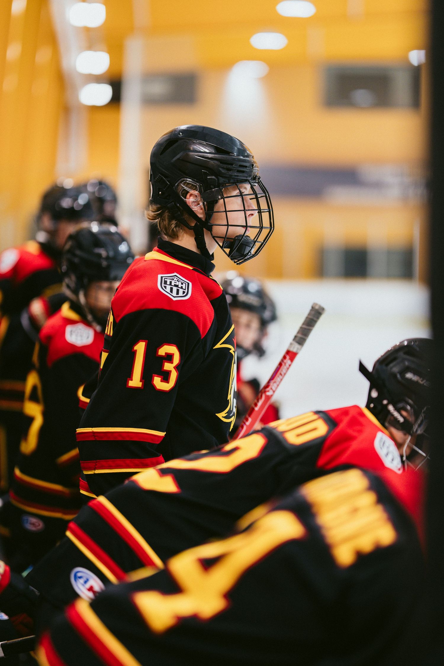 Hockey player wearing jersey number 13 sits on the bench during a game, wearing a black helmet with cage face guard.