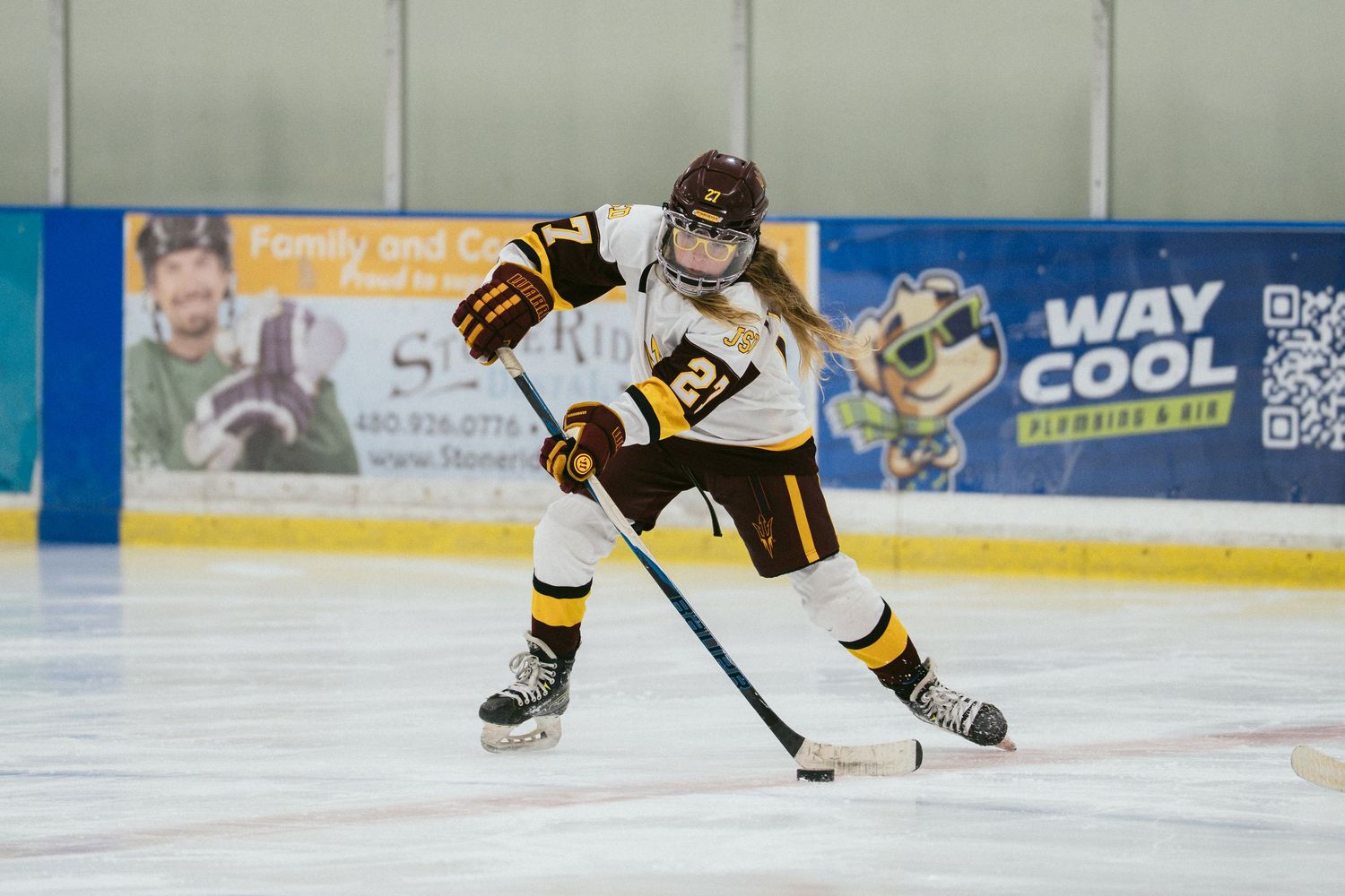Youth hockey player in white and gold uniform skating with puck on indoor ice rink during game.