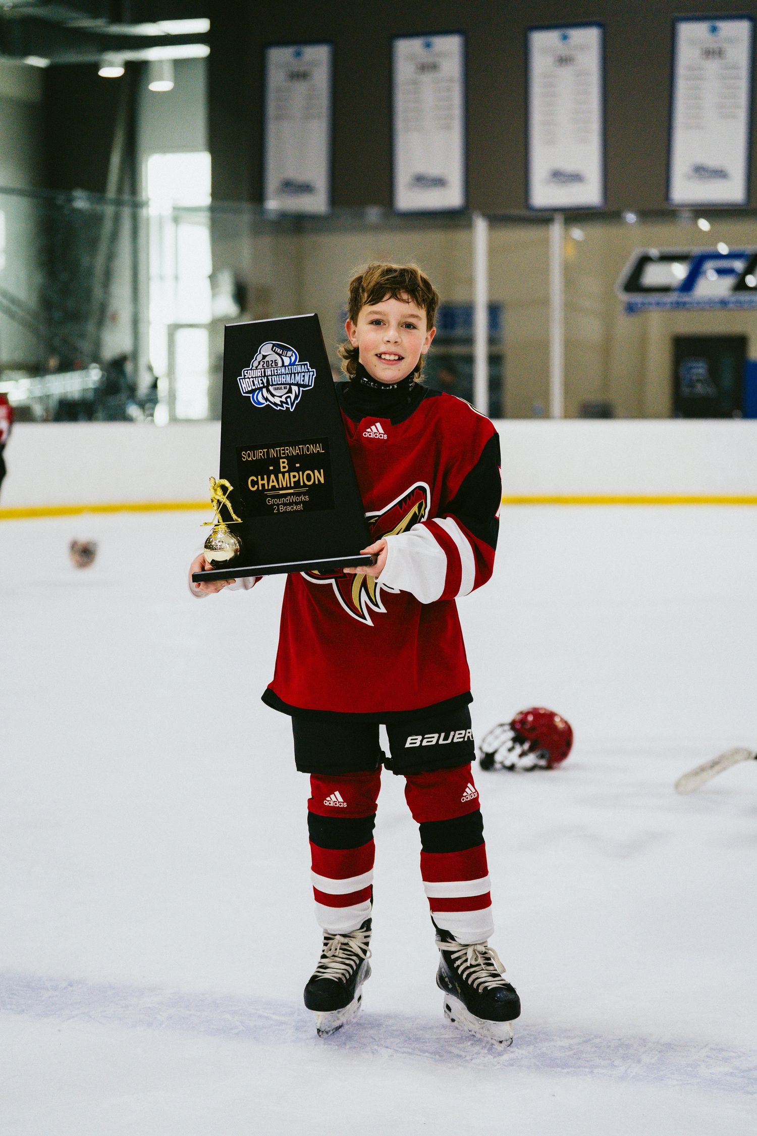 Small hockey player in red uniform holds championship plaque while skating on ice.