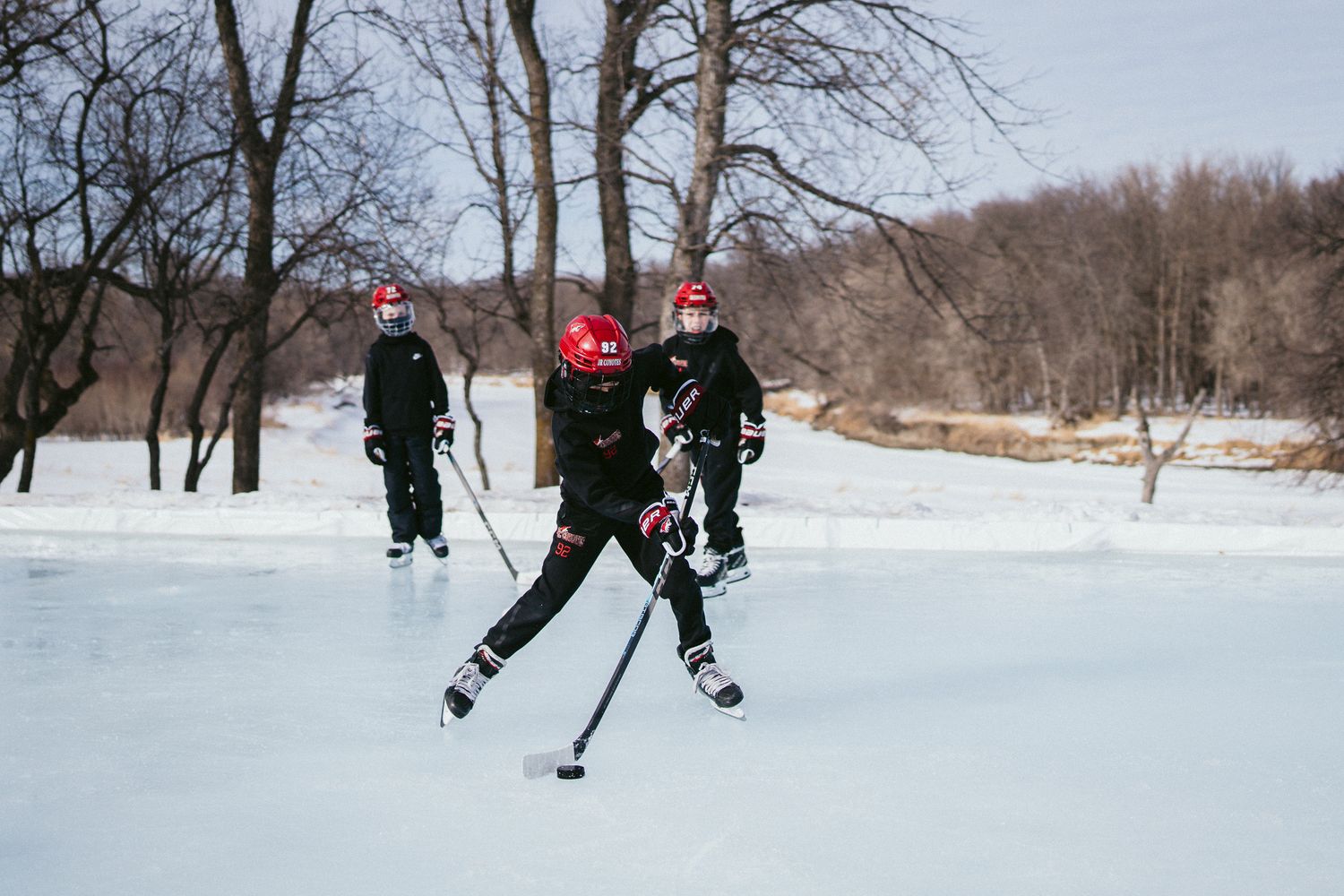 Hockey players skating on outdoor ice rink surrounded by snow-covered trees.