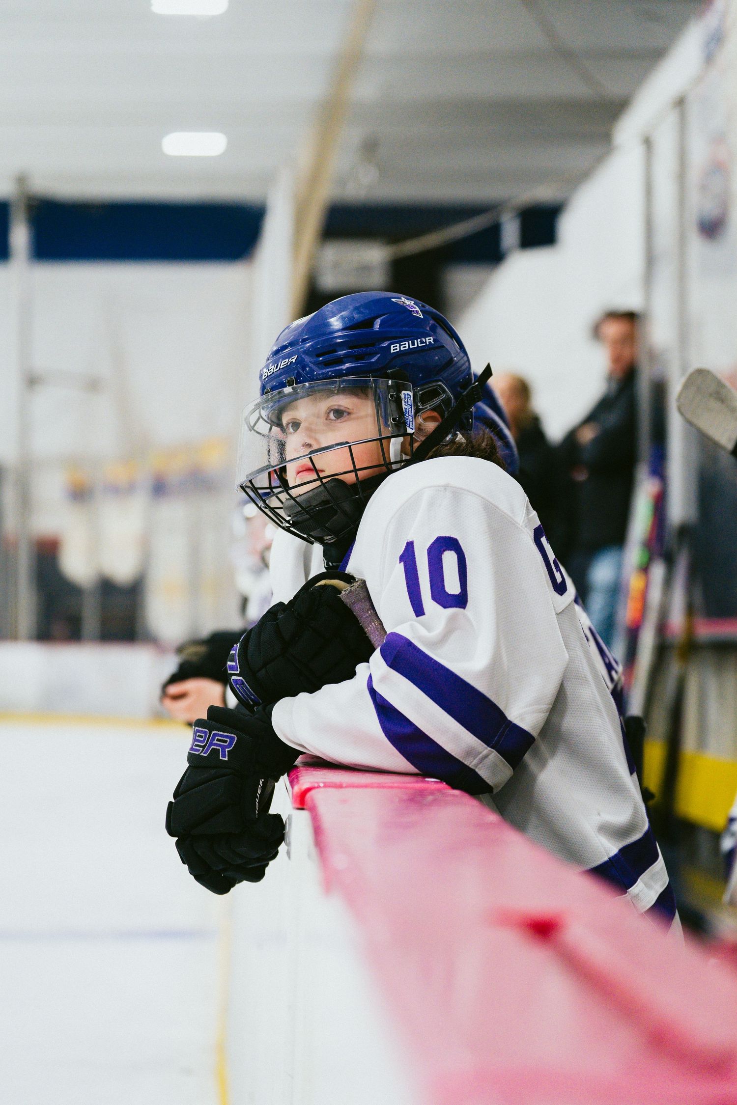 Young hockey player in blue helmet and white jersey leans against rink boards during game.
