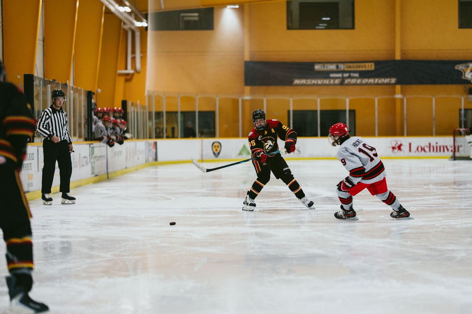 Two youth ice hockey players compete for the puck on an indoor rink as a referee watches nearby.