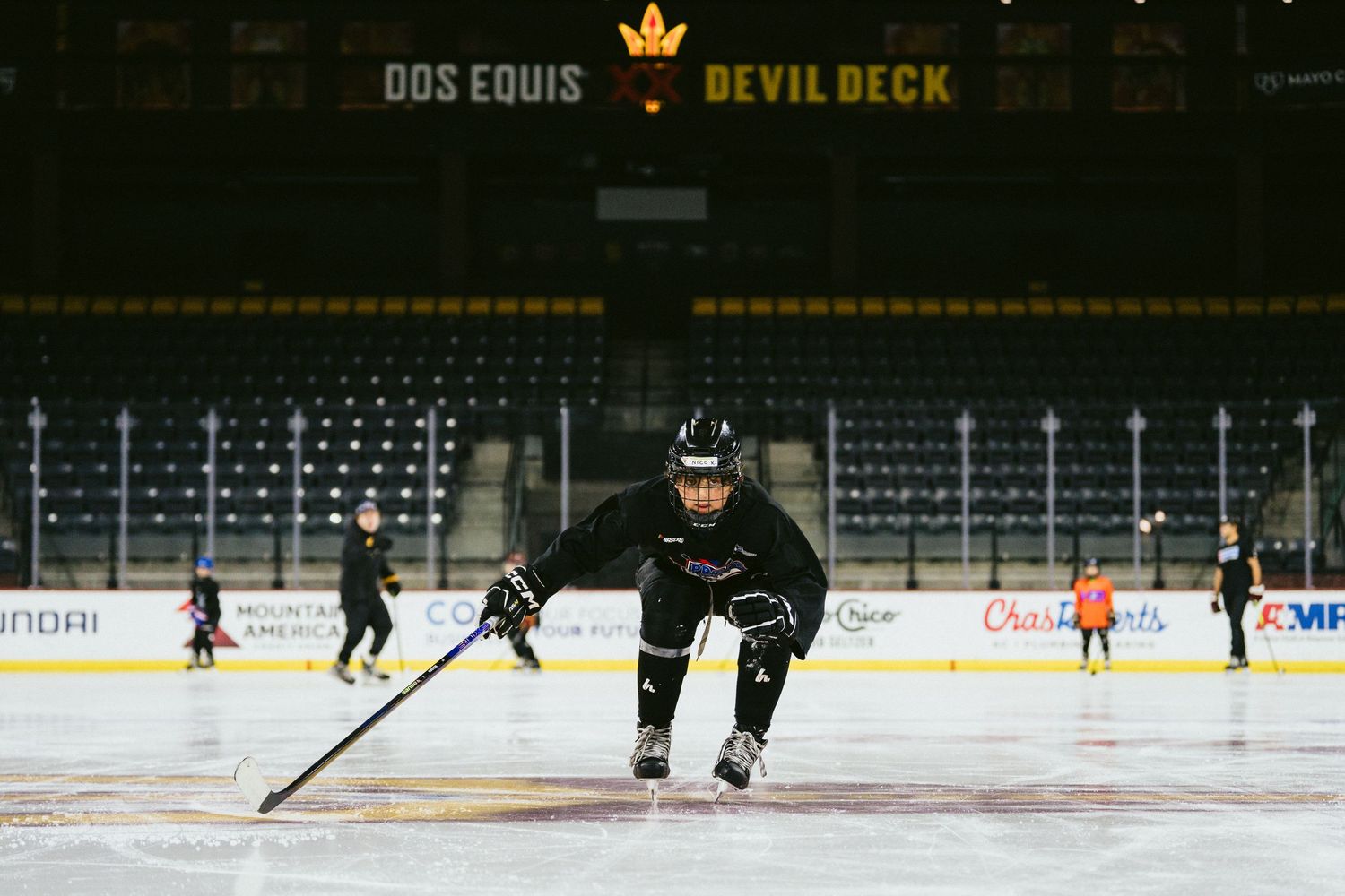 Hockey player in black uniform crouches during drill at ASU arena with Dos Equis Devil Deck sign visible.