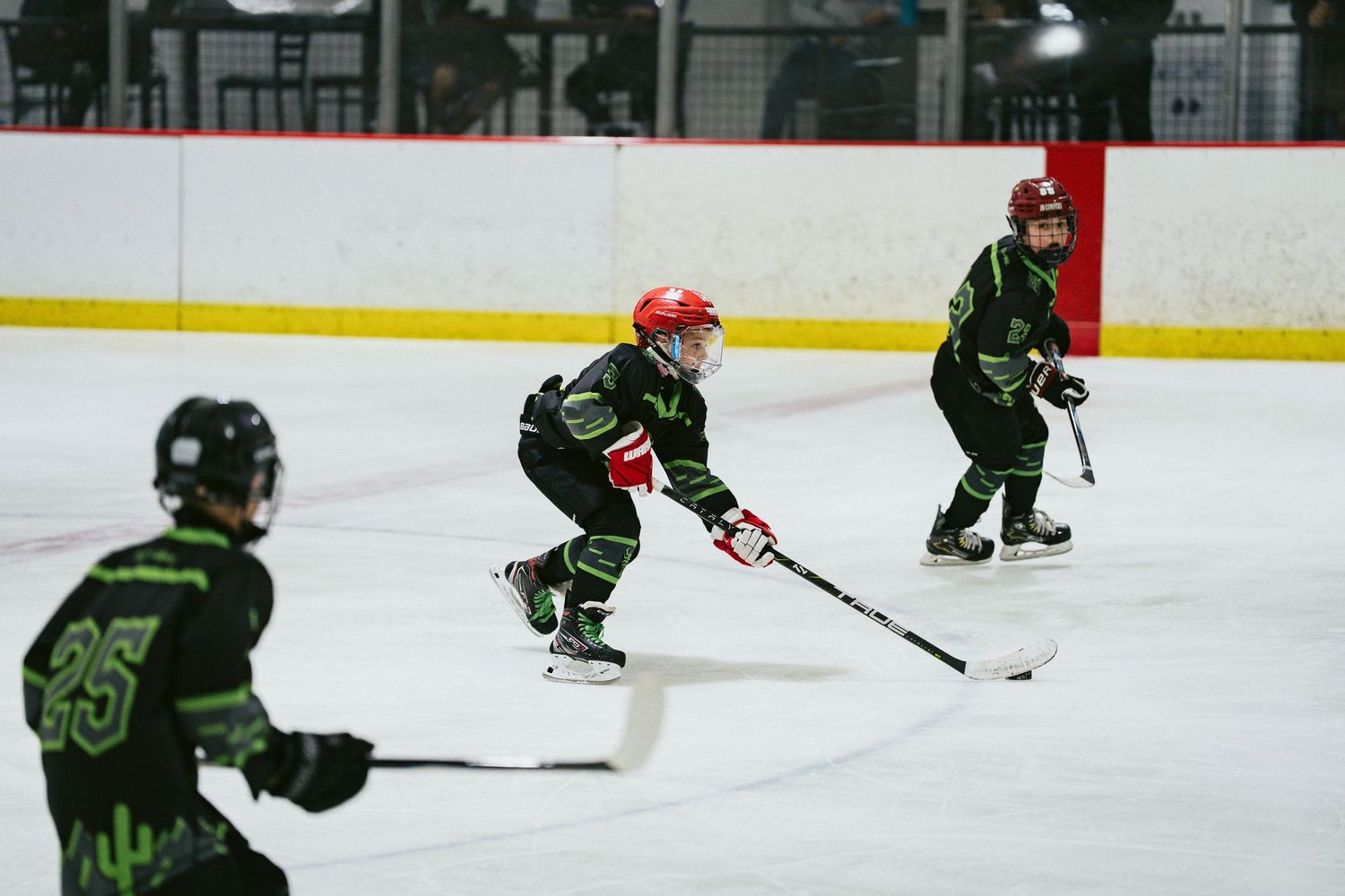 Three young hockey players in matching black and green uniforms skate during a youth ice hockey game.