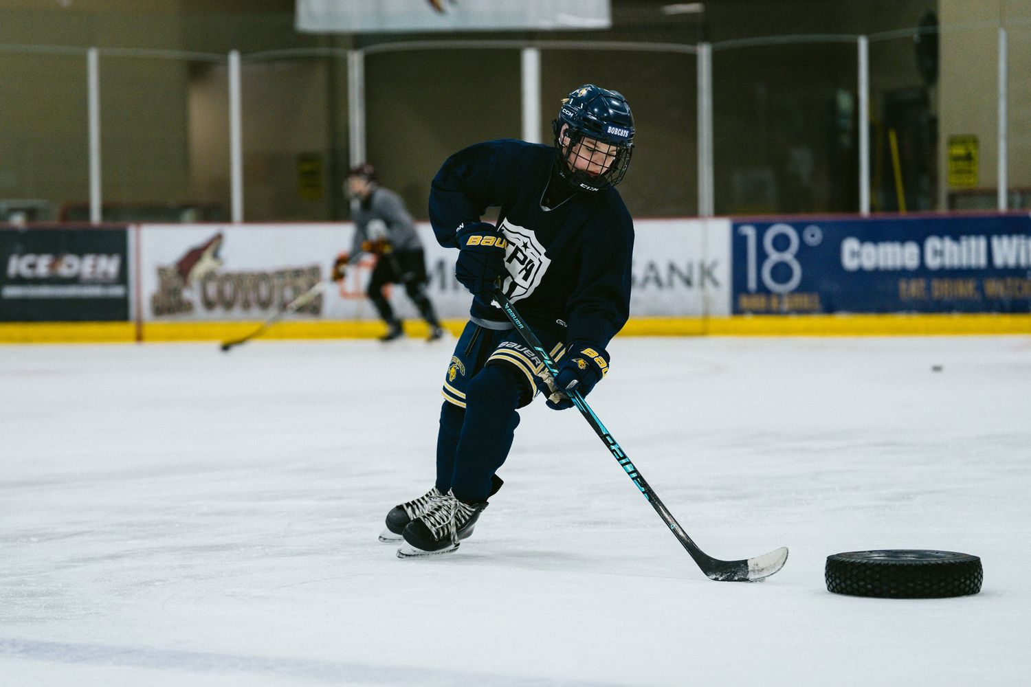 Young hockey player in dark uniform handles puck on ice during practice drill at indoor rink.