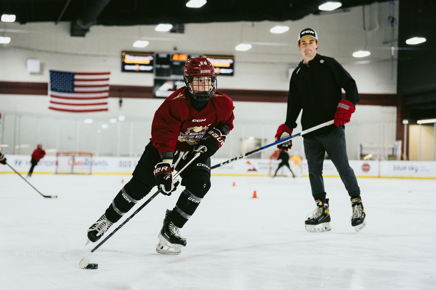 Youth hockey player in red jersey practicing puck handling on ice with coach observing during training session.