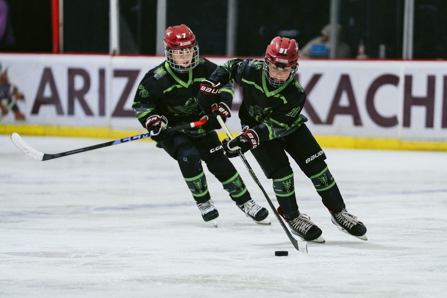 Two youth hockey players in black green uniforms skating fast during competitive ice hockey game.