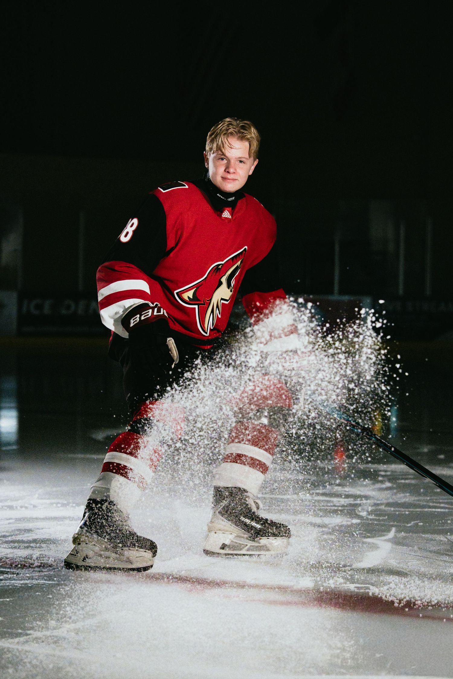 Hockey player in red Coyotes jersey stops on ice, spraying snow, during a dramatic arena photo shoot.
