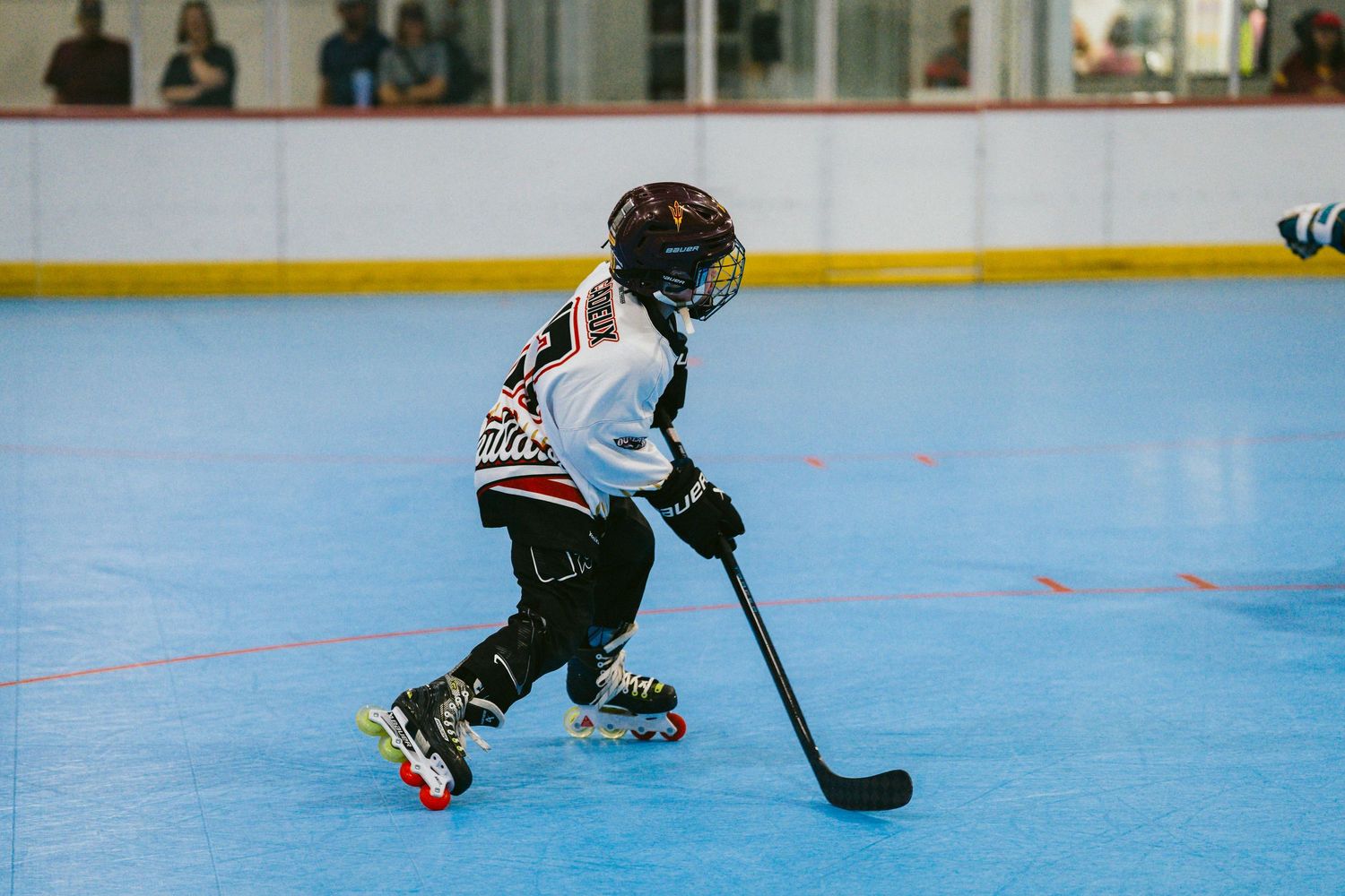 Young hockey player in white and black jersey skating with a stick on an indoor roller hockey rink.