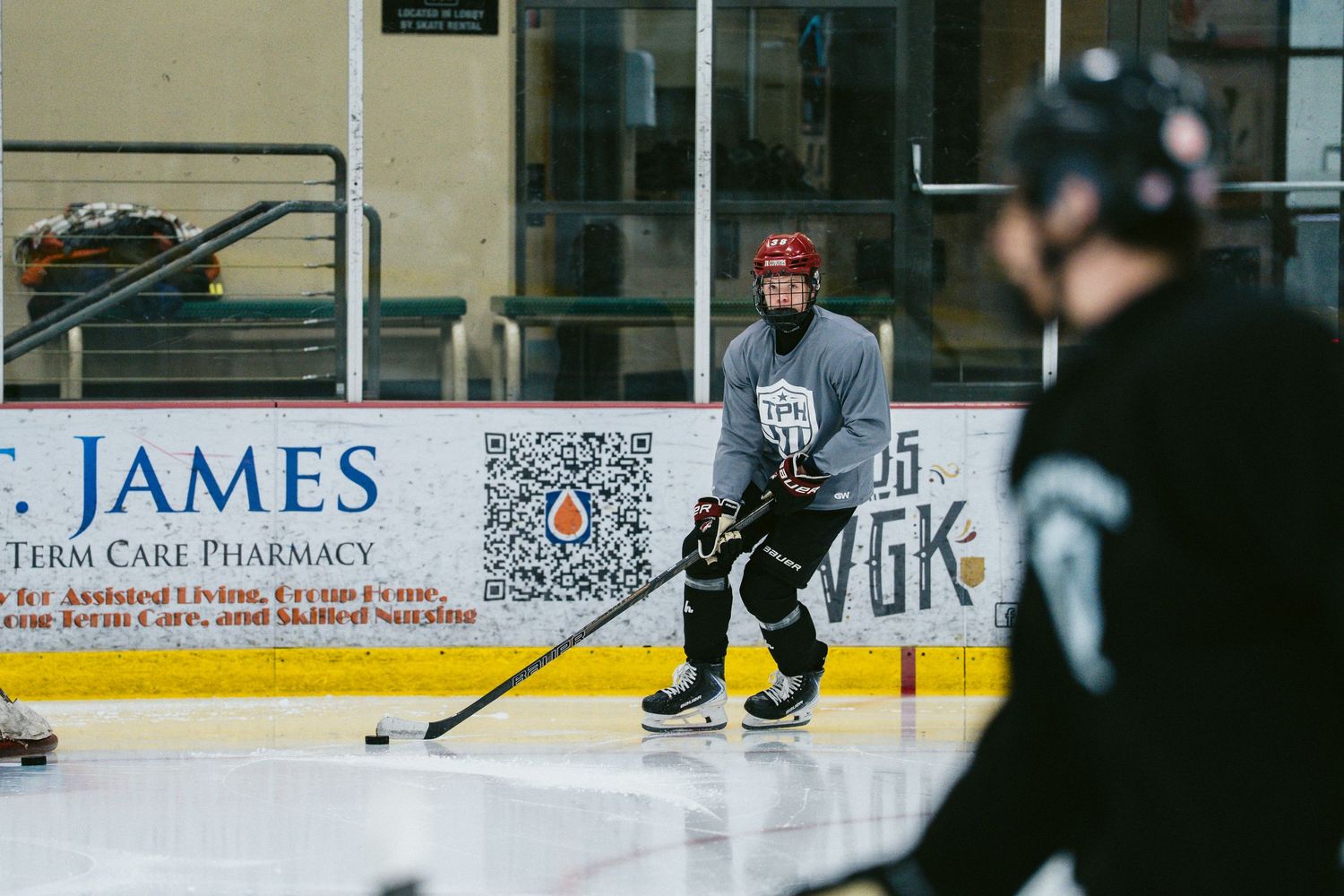 Hockey player in gray jersey handles puck near boards with St. James Pharmacy advertisement during practice.