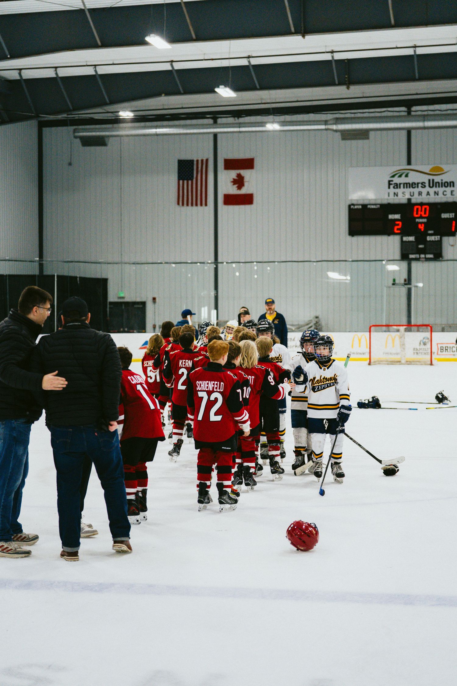 Youth hockey teams line up on ice rink with American and Canadian flags visible on wall.