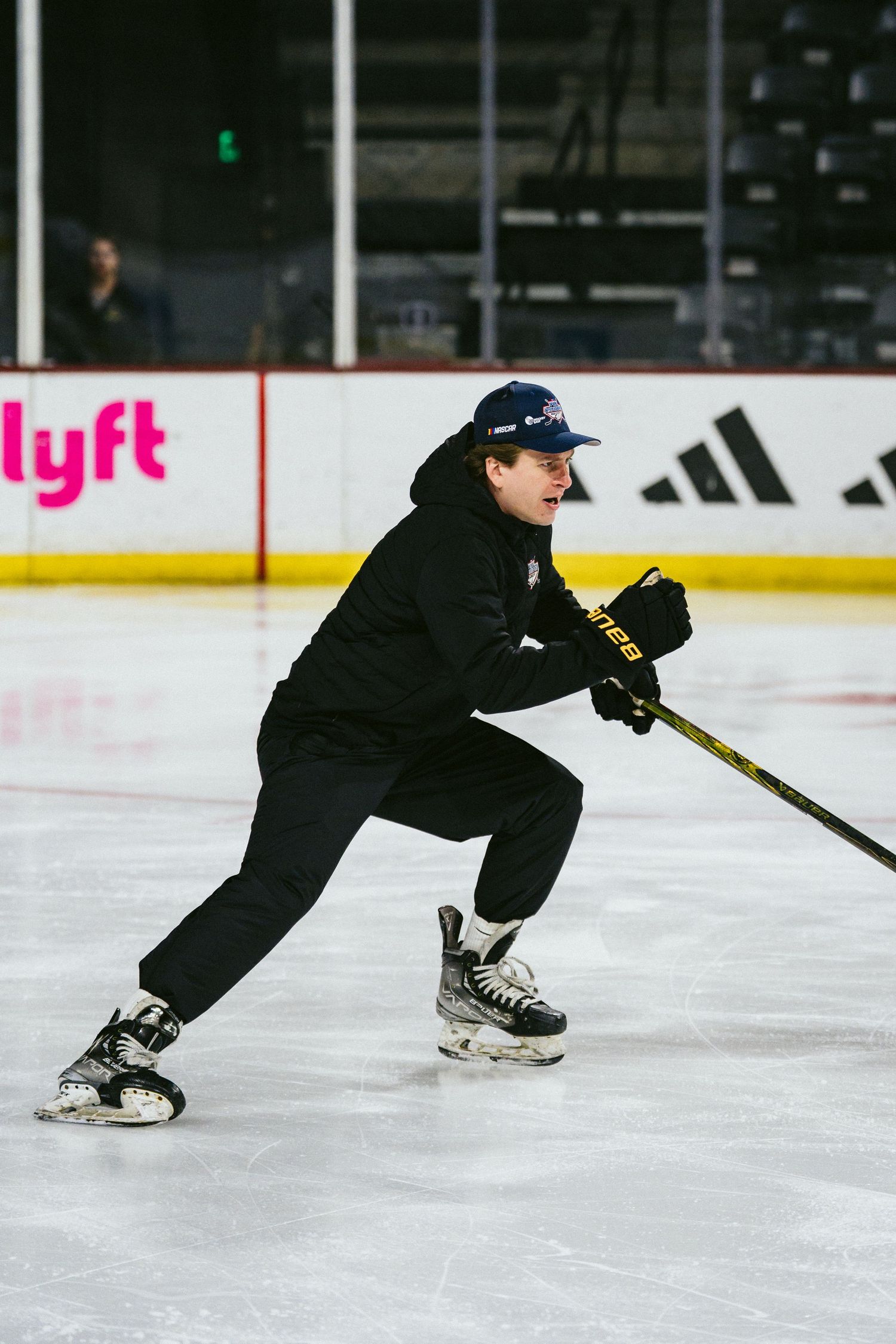 Hockey player in black outfit and cap skating with a Bauer stick on an indoor ice rink.