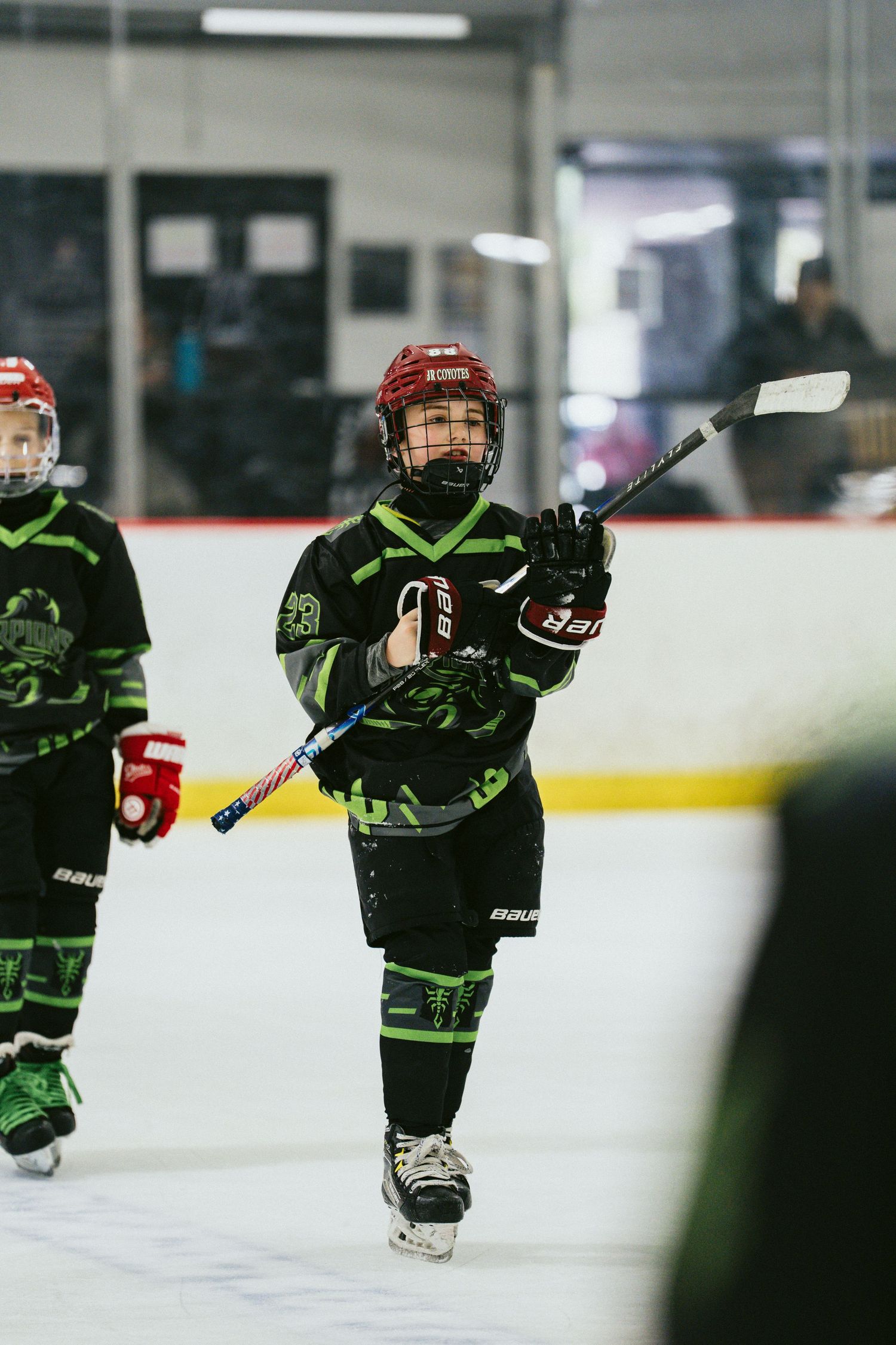 Youth hockey player in black green uniform raises stick while skating on indoor ice rink.