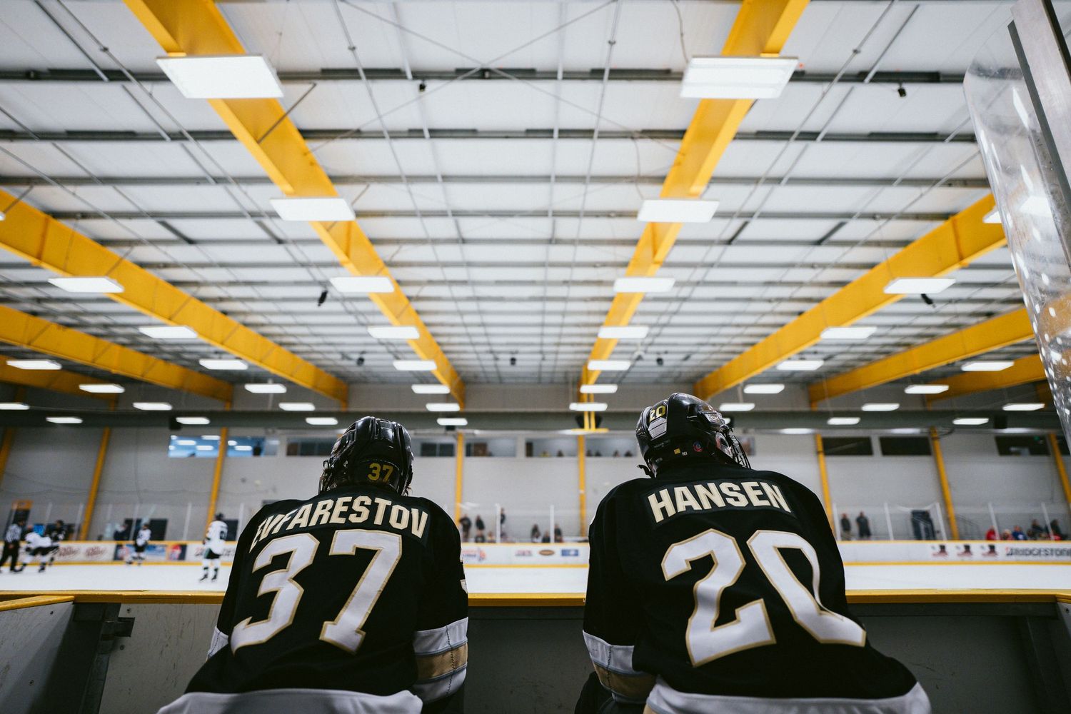 Two hockey players wearing jerseys numbered 37 and 20 sit on the bench inside an indoor ice rink.