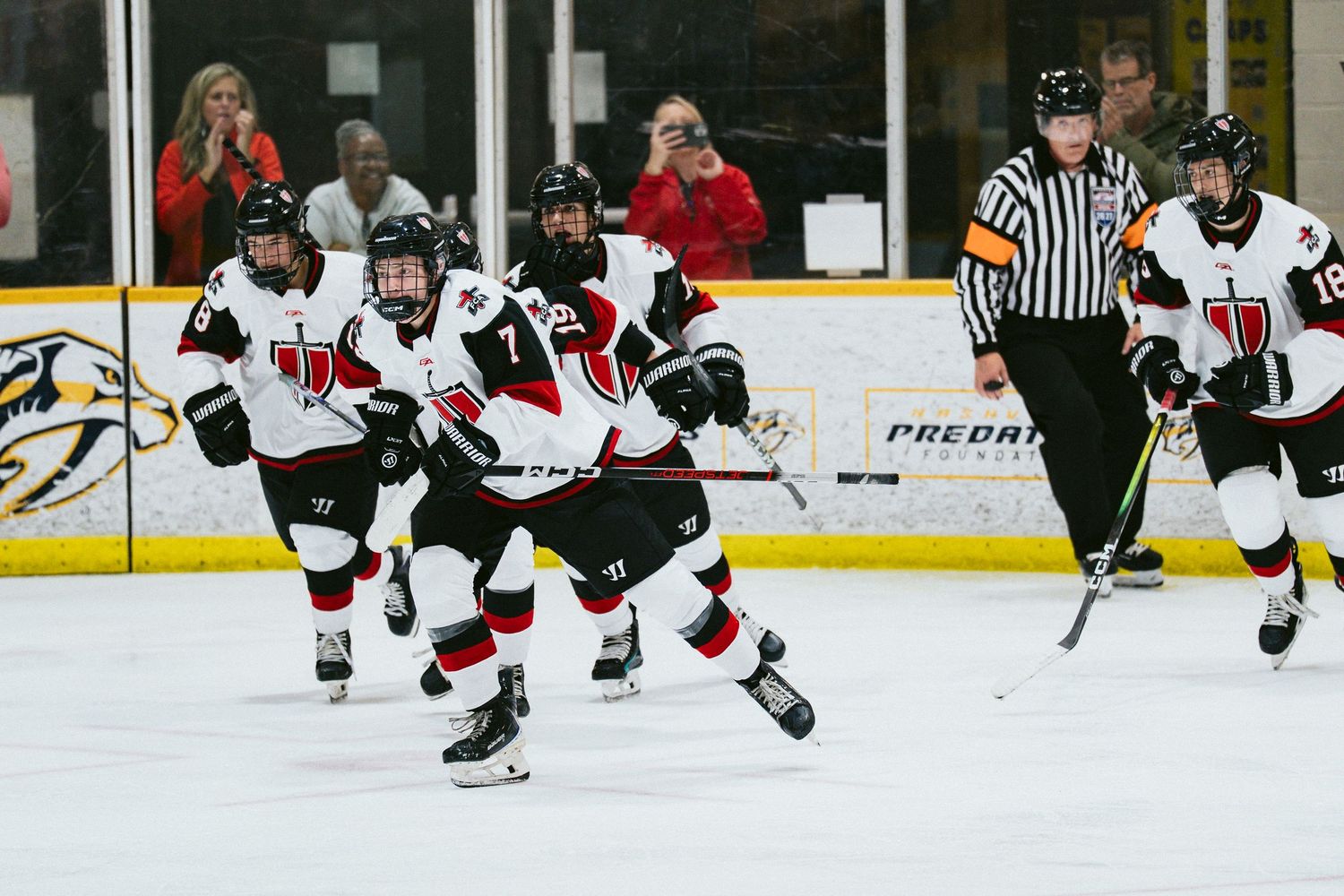 Youth hockey players in red and white uniforms skate on indoor ice rink during competitive game with referee nearby.