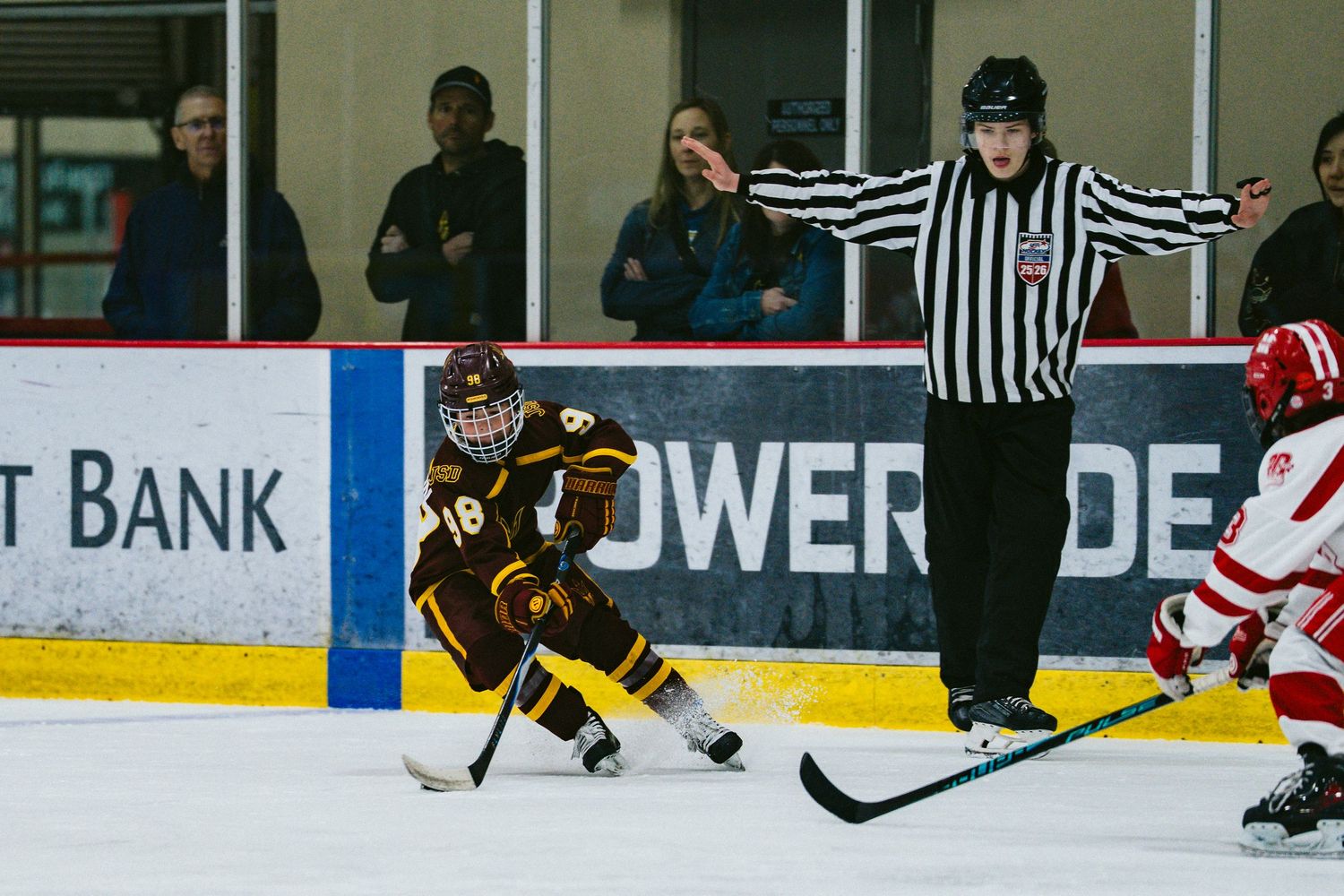 Hockey game officials in striped uniforms oversee match between teams in brown and red jerseys.