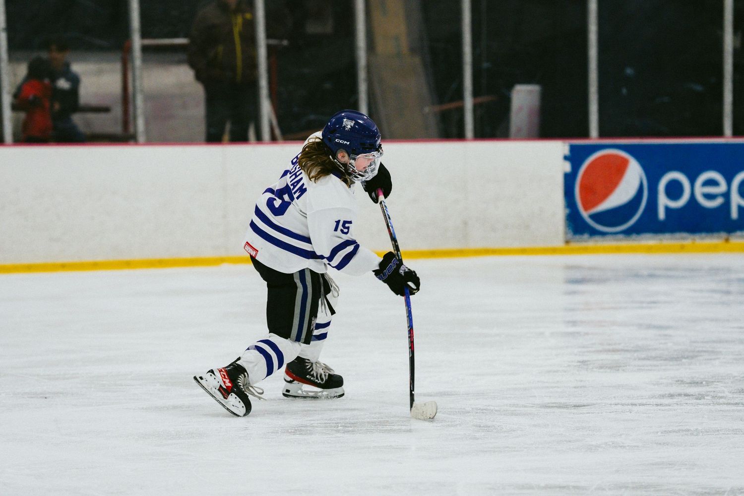 Youth hockey player skating with stick on ice in white and blue uniform during game action.