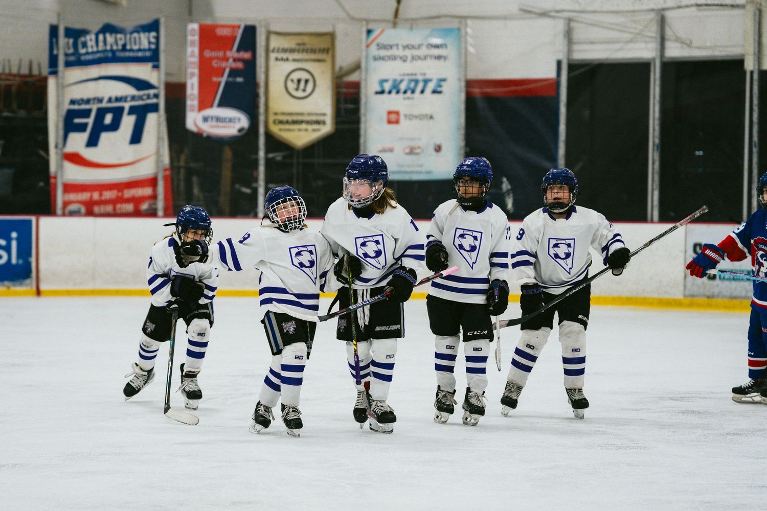 Youth hockey team in blue and white jerseys lines up along the rink boards.