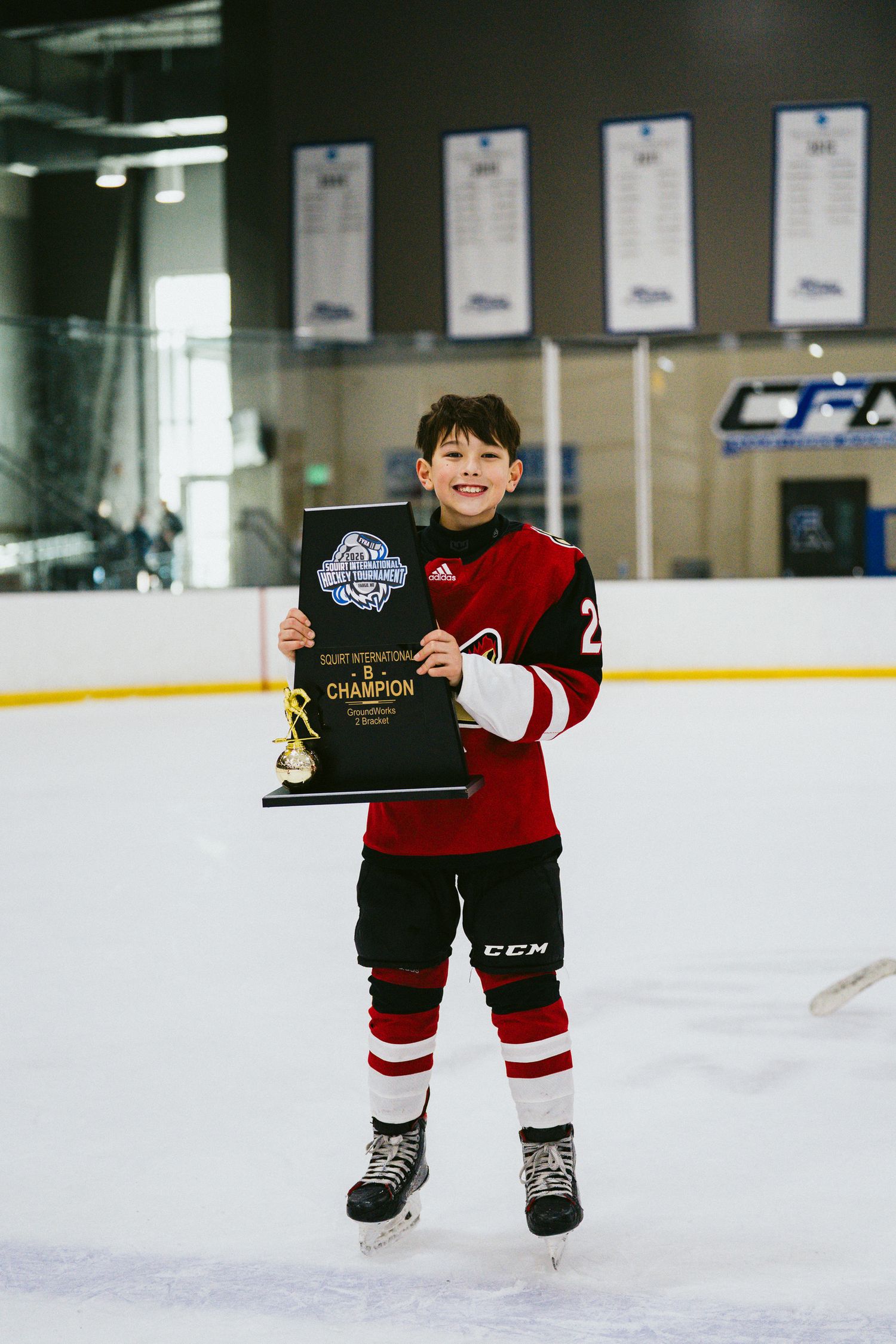 Small hockey player in red and black uniform displays award on ice.
