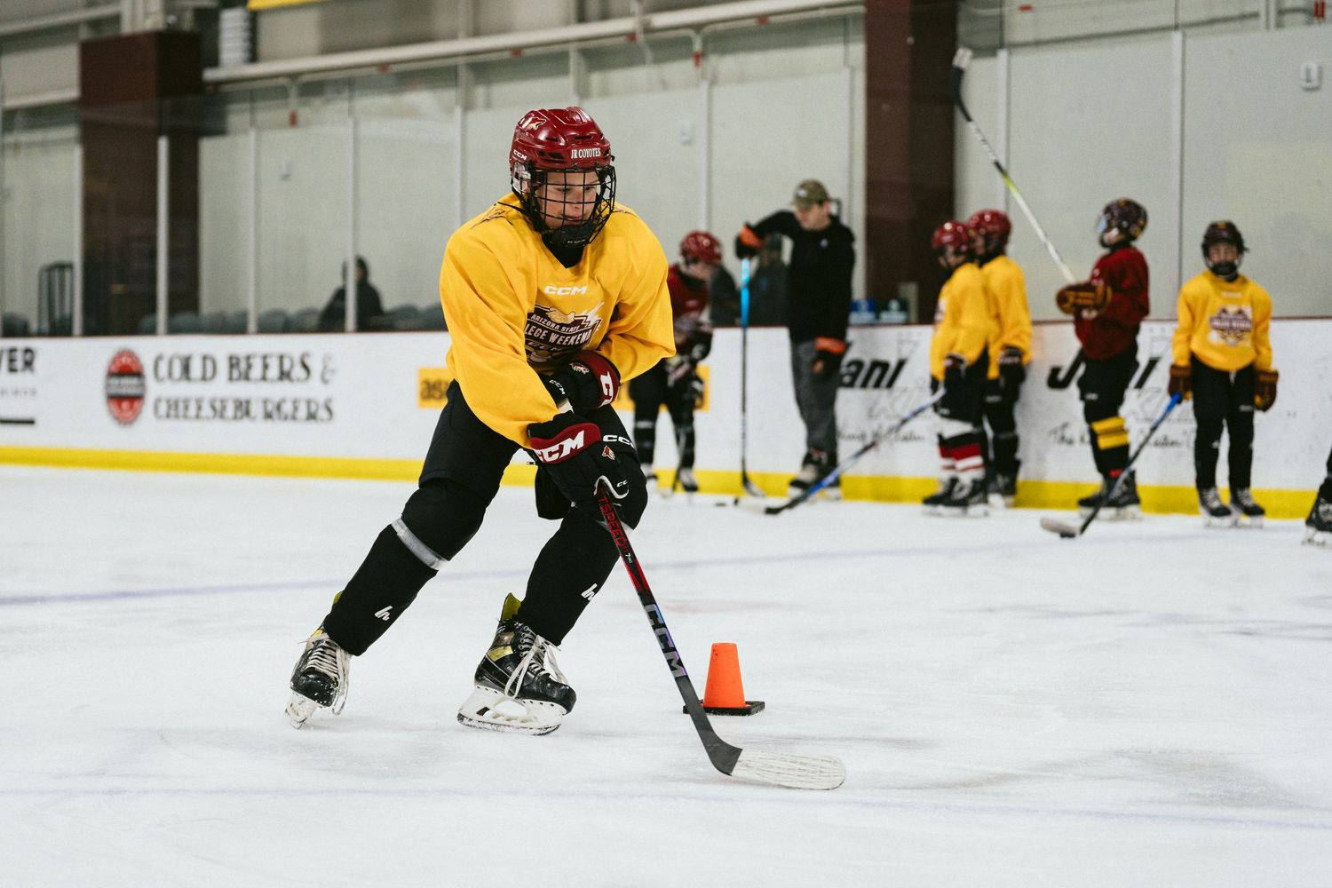 Young hockey player in yellow jersey maneuvering puck around cone during ice practice.
