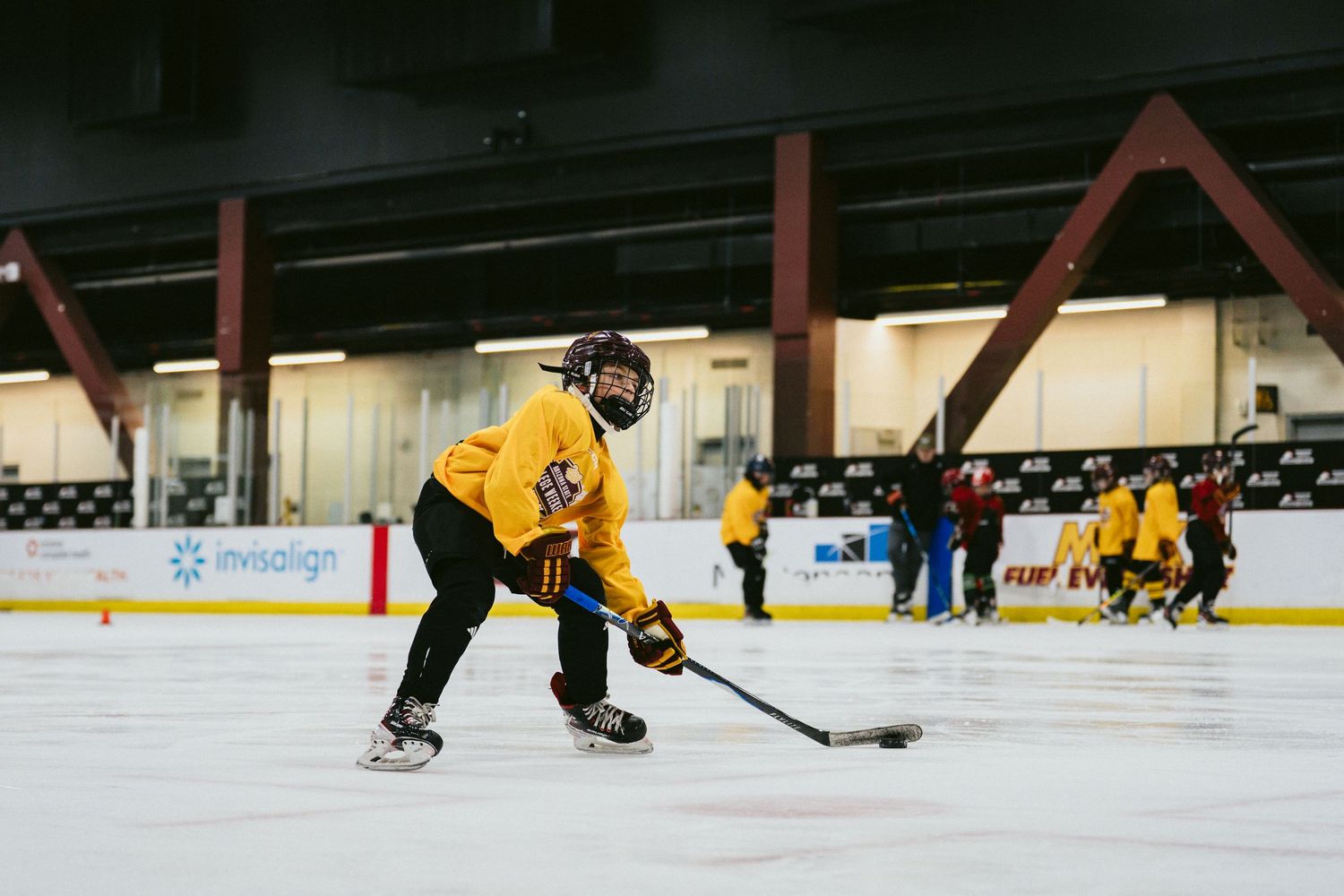Youth hockey player in yellow uniform skates with puck during indoor ice rink training session.