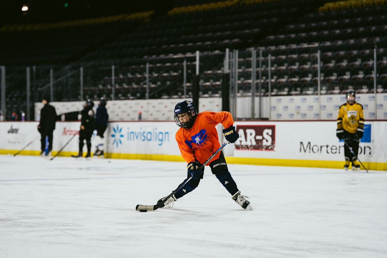 Youth hockey player in orange jersey skating with puck on indoor ice rink during practice.