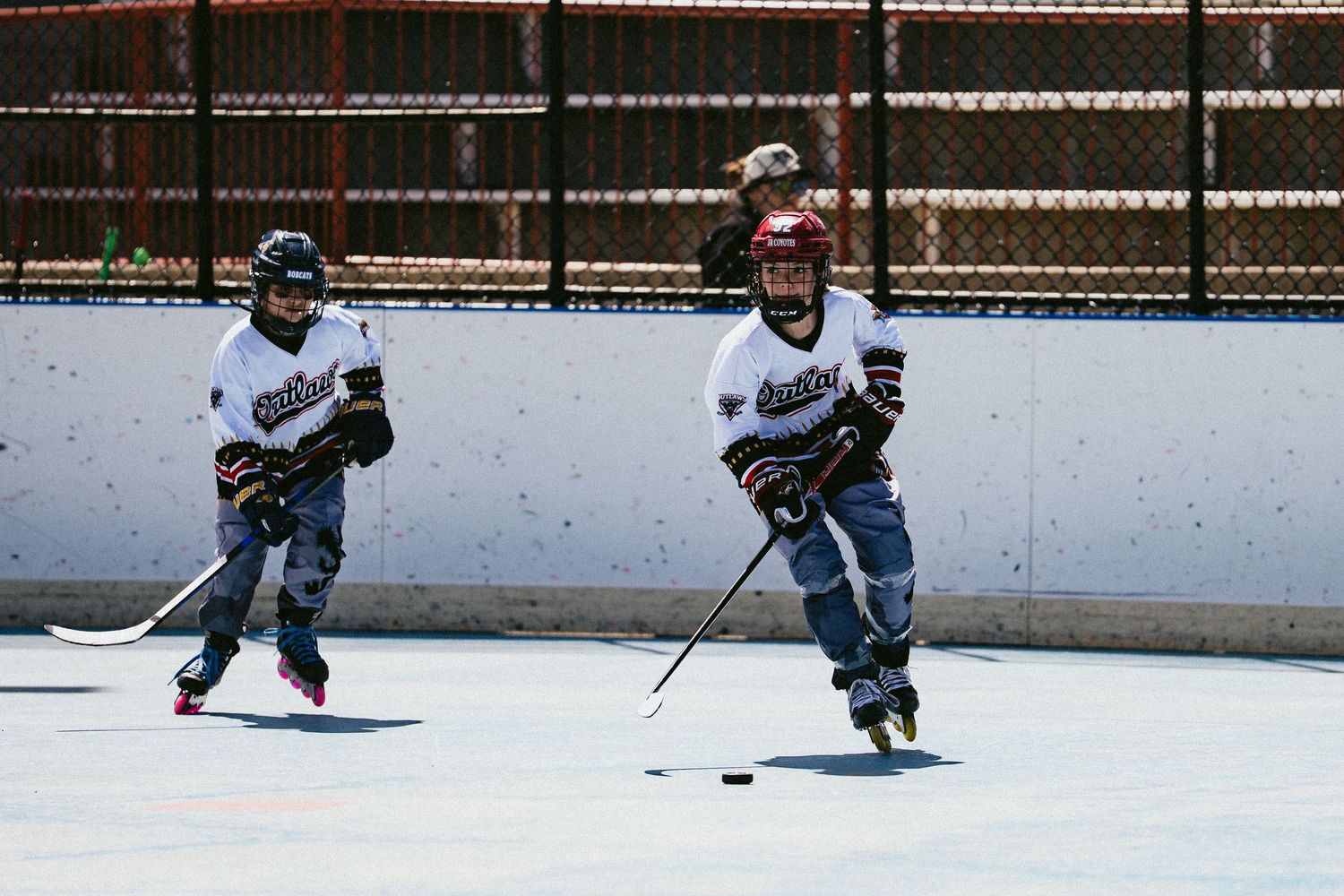 Young children in hockey gear play roller hockey on an outdoor rink during a sunny day.