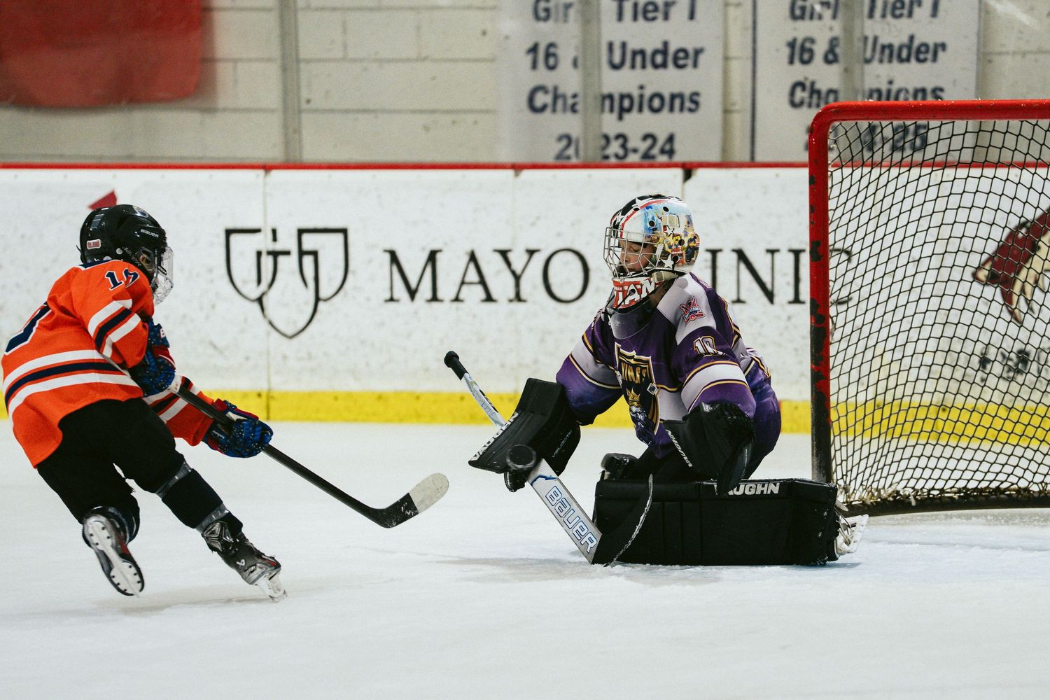 Youth hockey goalie in purple makes save against orange team player during competitive ice hockey game.
