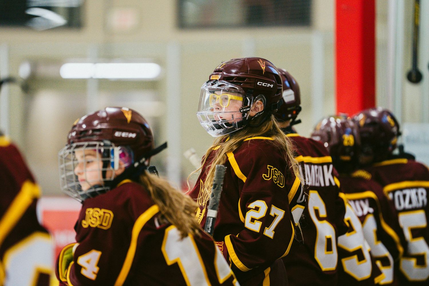 Youth hockey players in brown and gold jerseys watch intently from the bench during a game.
