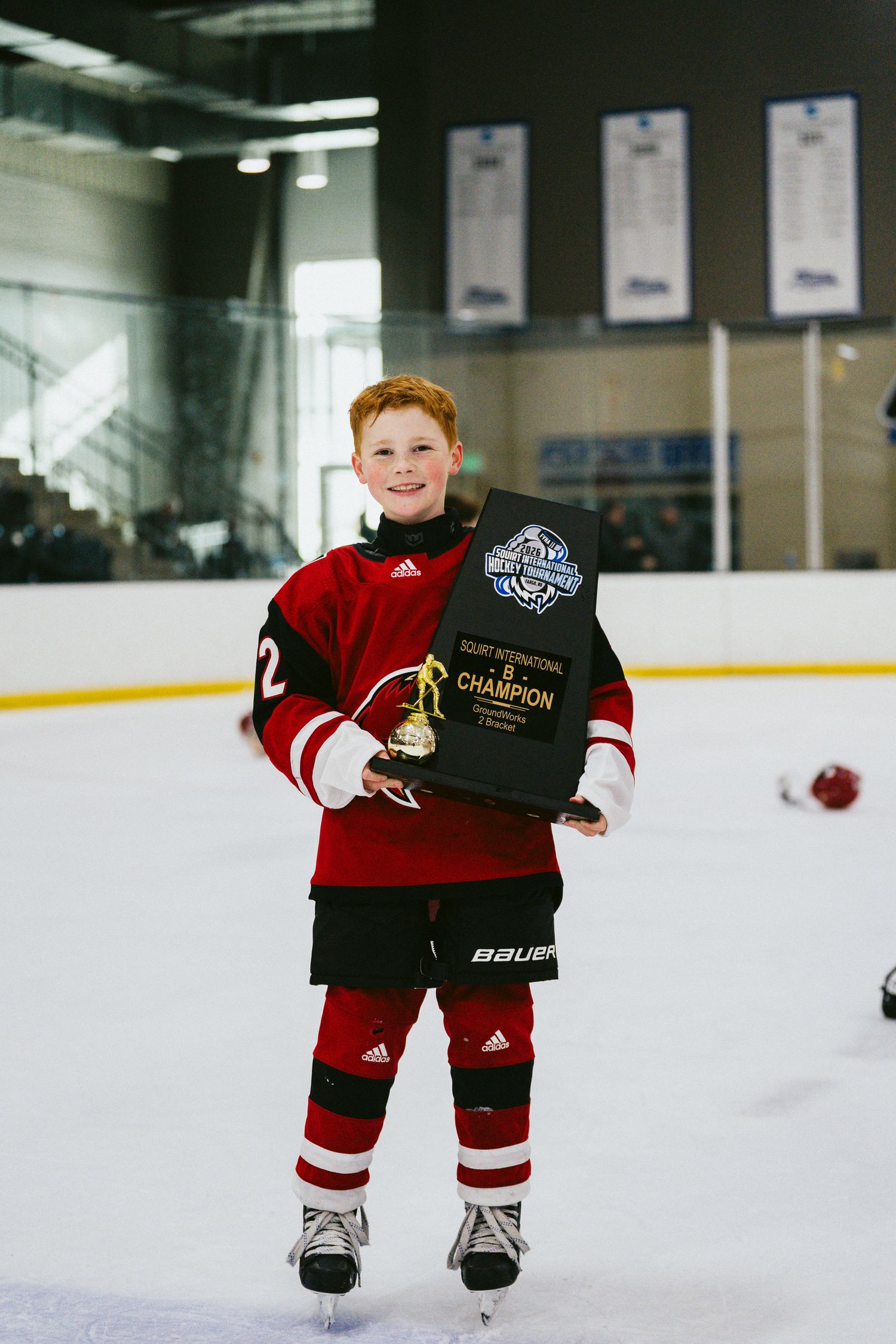 Young hockey player in red uniform posing with championship trophy on ice.
