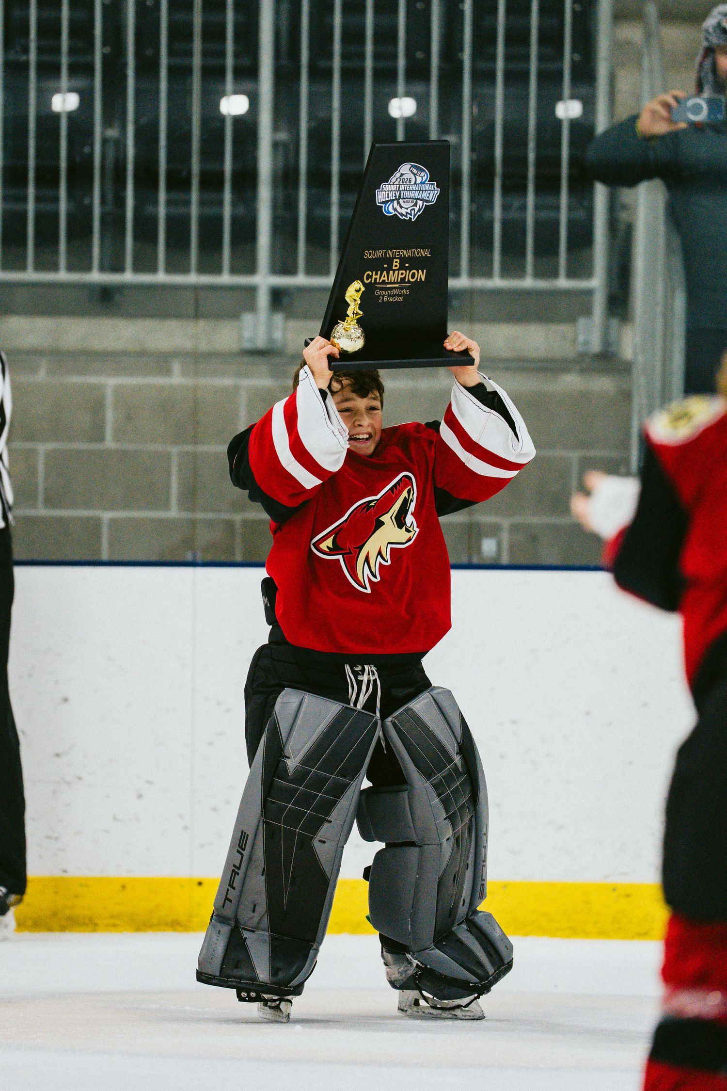 Hockey goalie in red jersey and pads stretching on ice during practice.