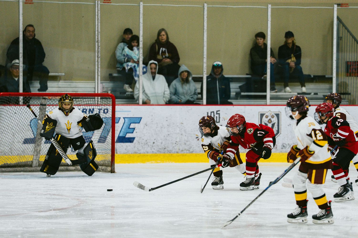 Youth hockey players in red and yellow jerseys compete during an ice hockey game at an indoor rink.